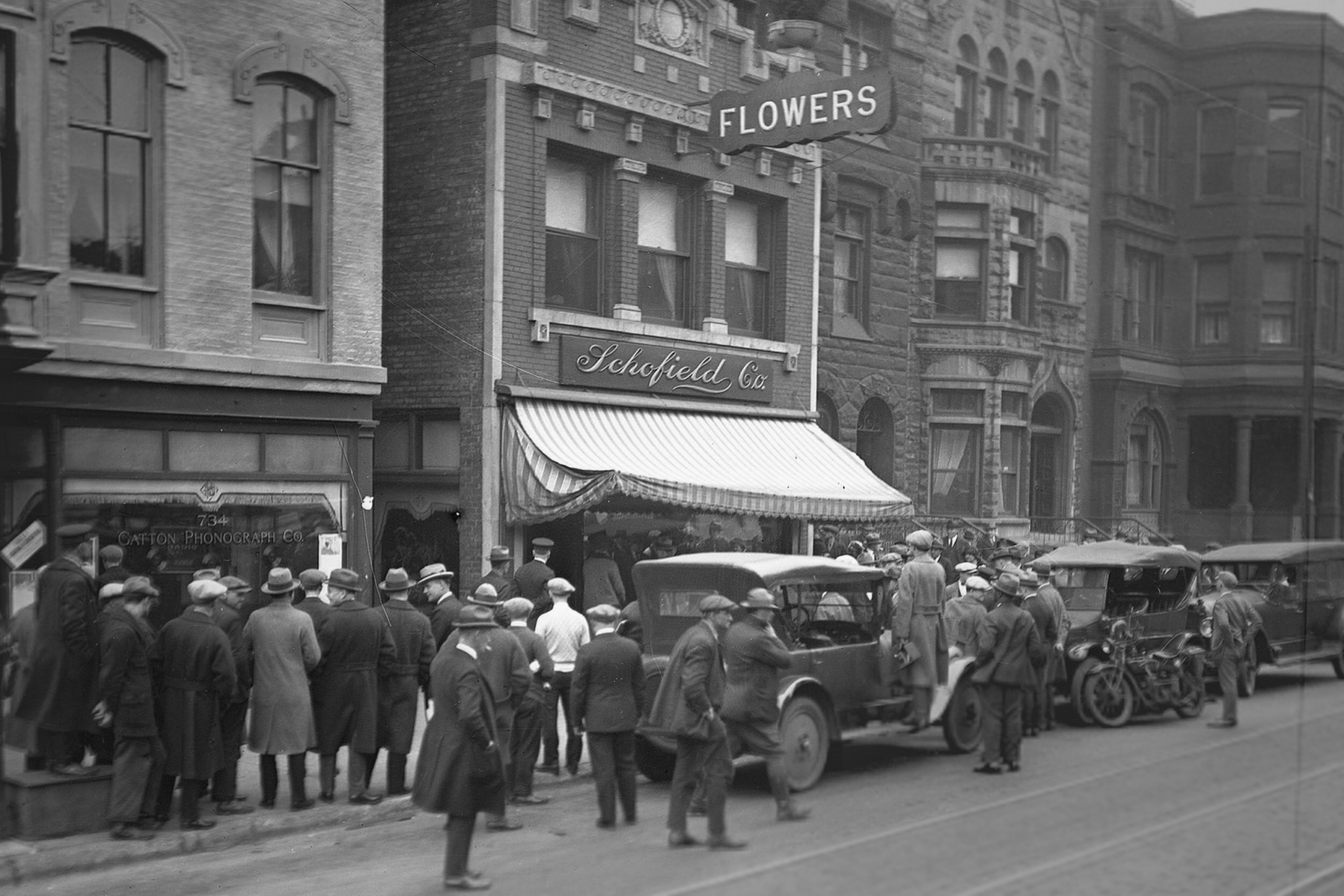 Group of people and vintage cars on a city street in front of buildings with storefronts and signs.