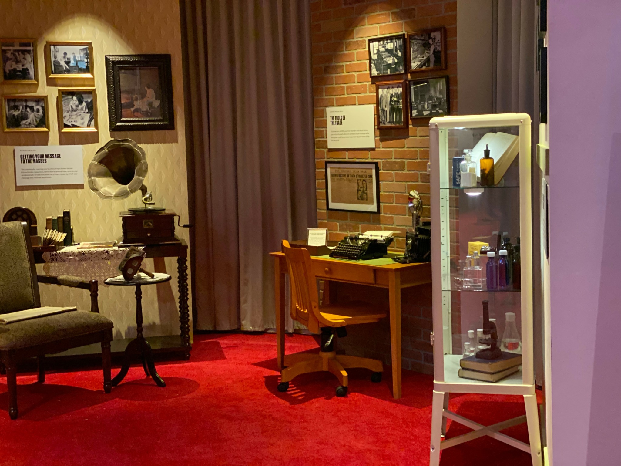 Room with framed pictures, a desk with a chair, a display cabinet, and a red carpet.