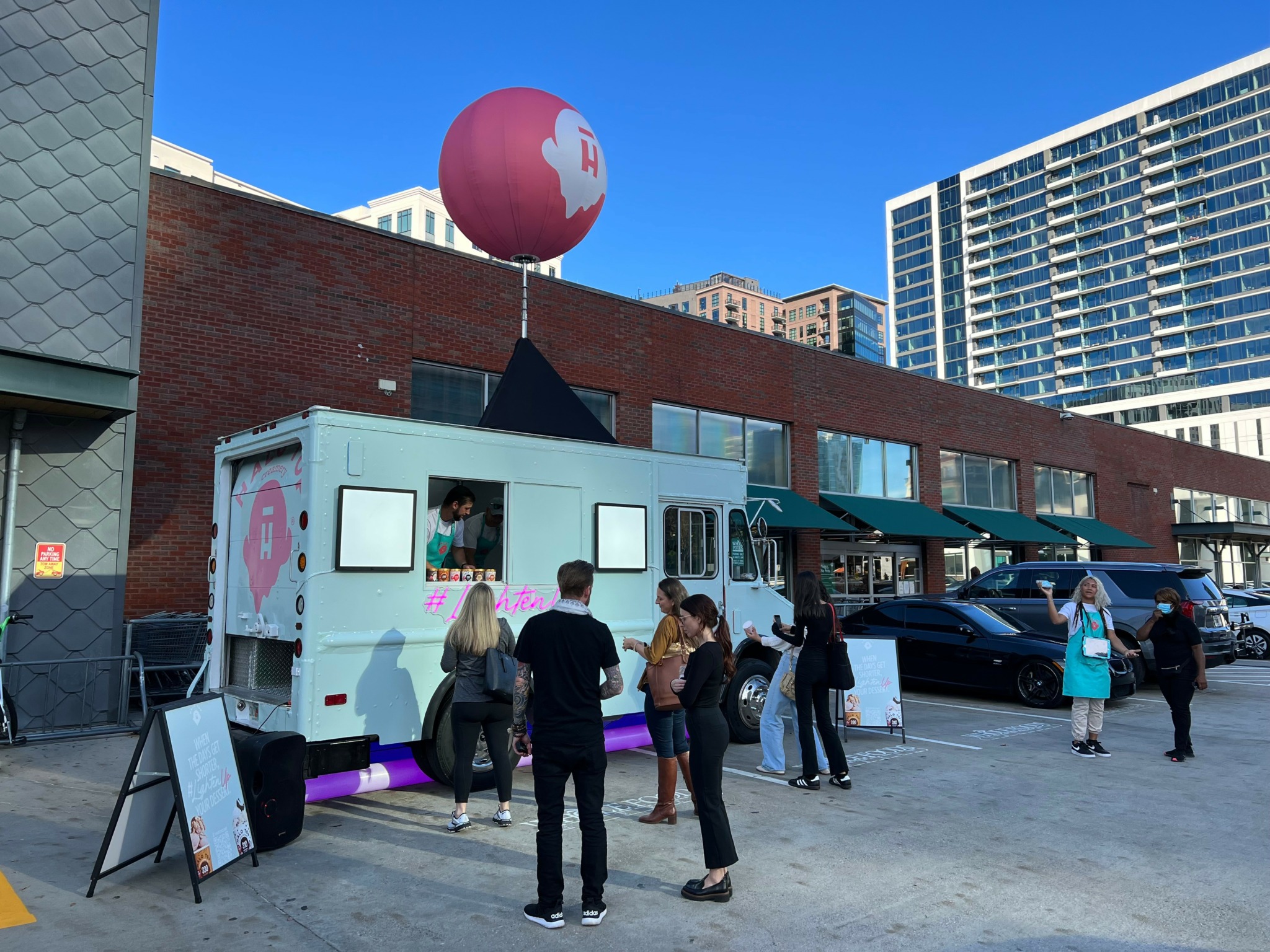 People gather around a white food truck with a pink balloon on top, parked in an urban area with buildings.