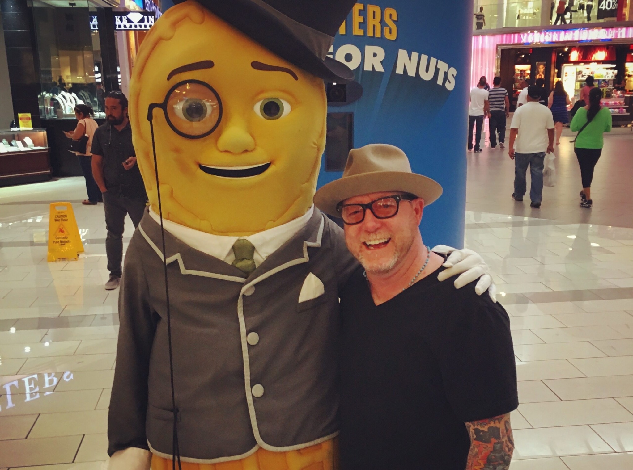 Person with glasses and hat smiling next to a large yellow mascot with glasses and a suit in a shopping mall.