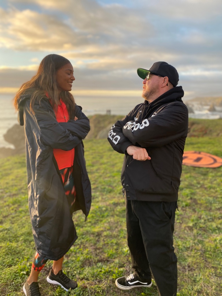 Two people standing outdoors on grass, facing each other and talking, with a cloudy sky in the background.
