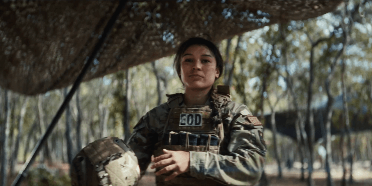 Female soldier in camouflage uniform holding a cup outdoors under a shaded canopy.