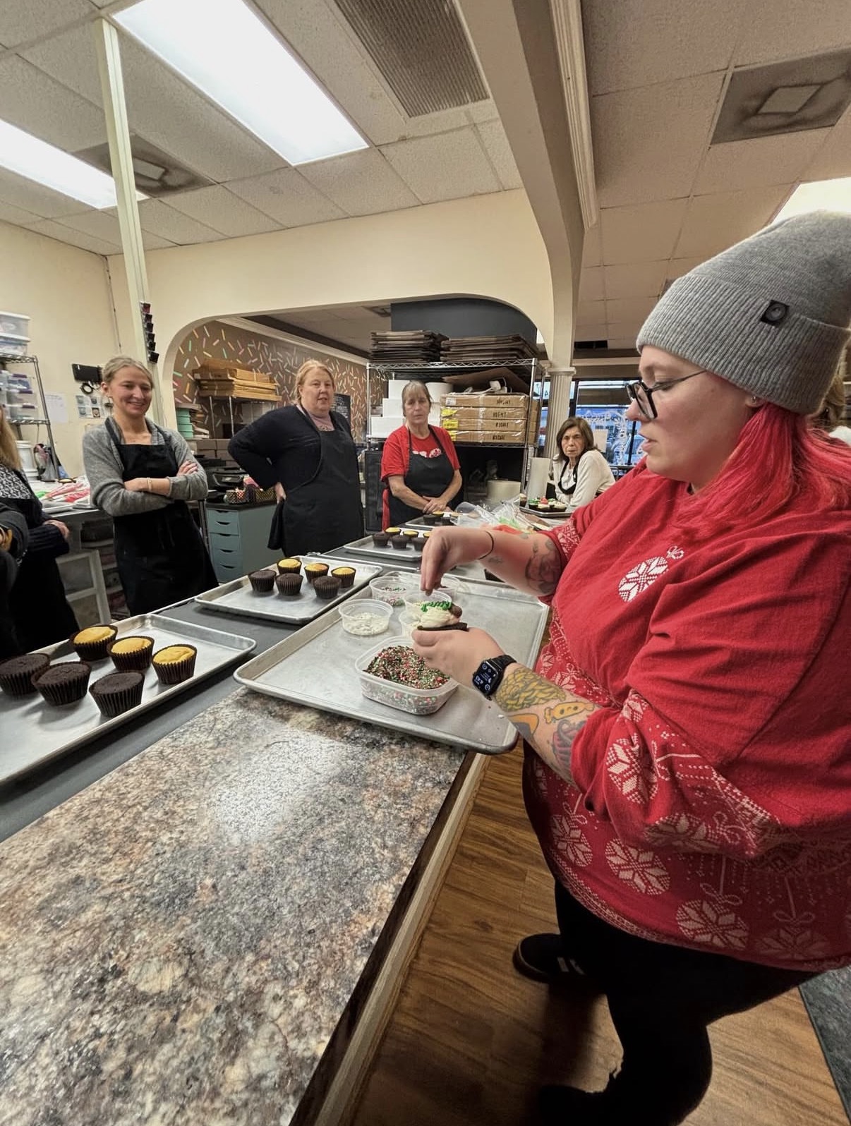 Woman with red hair and gray beanie preparing food at a counter in a kitchen or bakery, with people watching.
