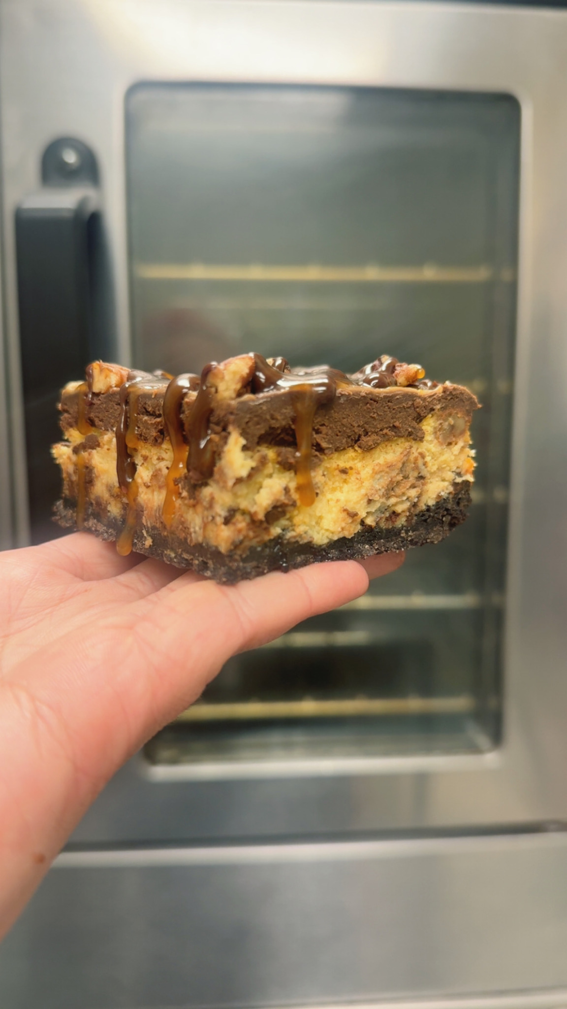 Hand holding a large cookie with chocolate chips and a thick chocolate coating, in front of a metallic oven.
