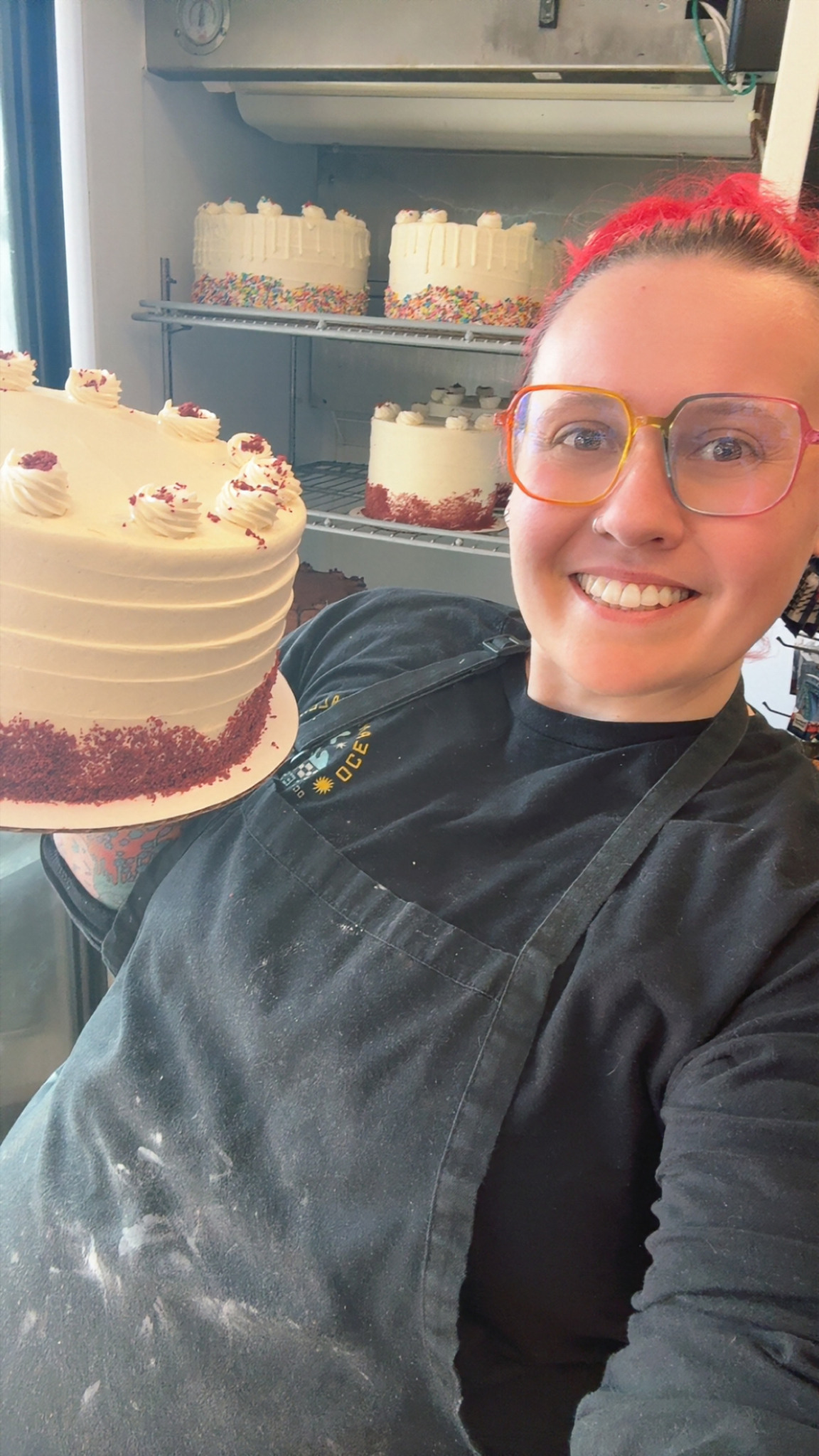 Person with red hair and glasses holding a decorated cake in a bakery kitchen.
