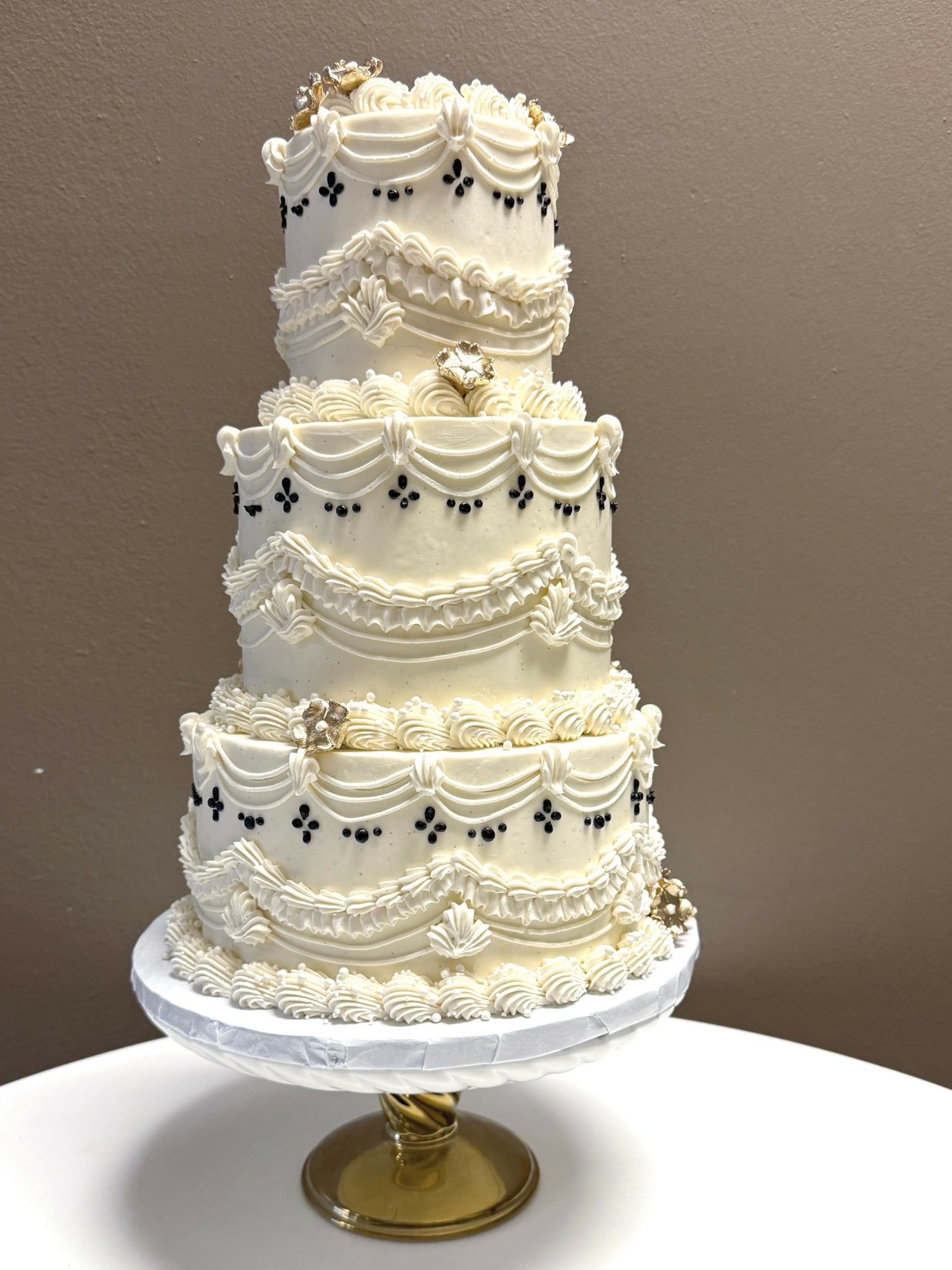 Four-tiered white wedding cake with decorative piping, gold accents, and small black and gold embellishments, on a gold stand.