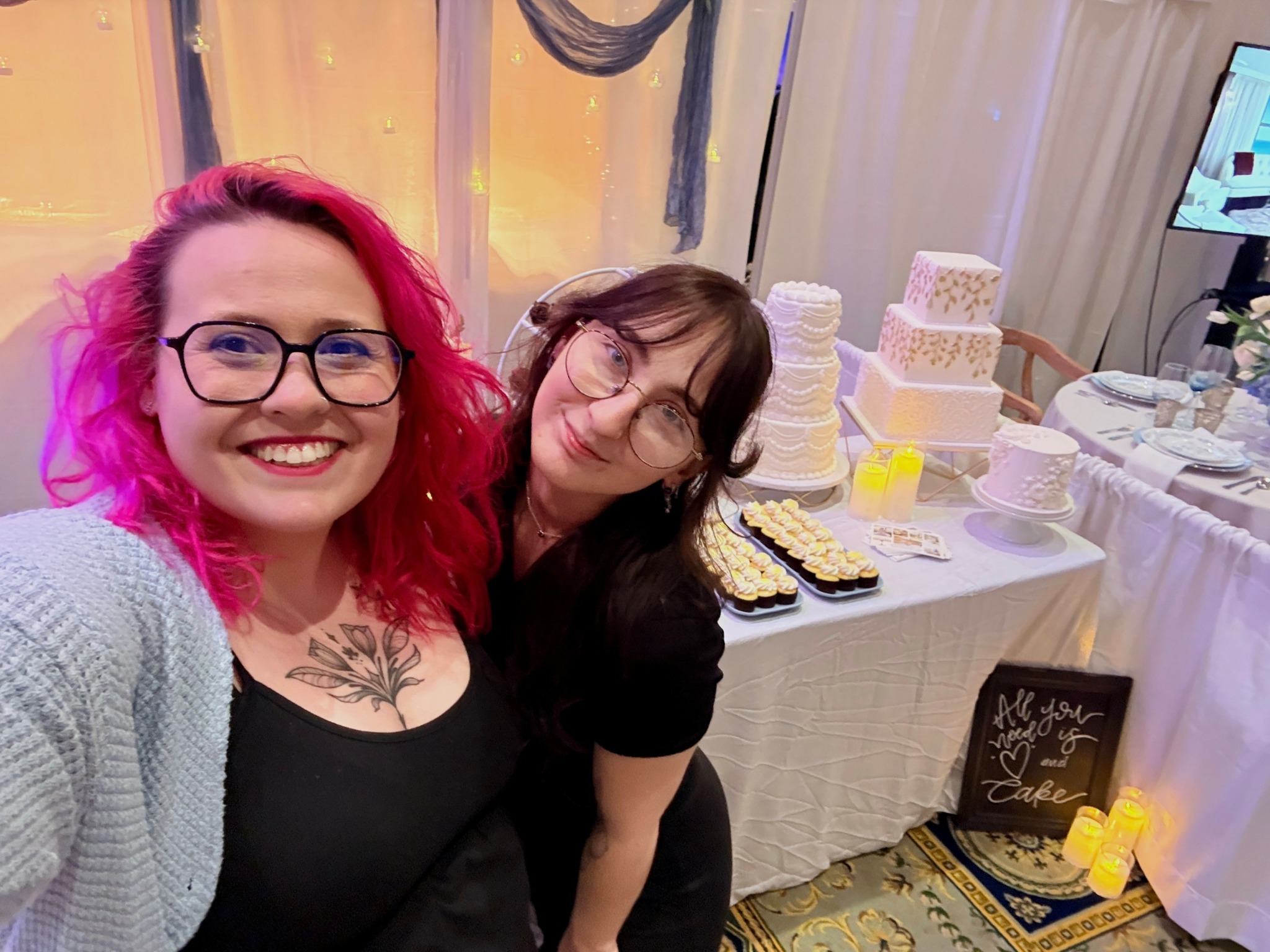 Two women smiling, one with pink hair and glasses, the other with brown hair, in front of a dessert table.