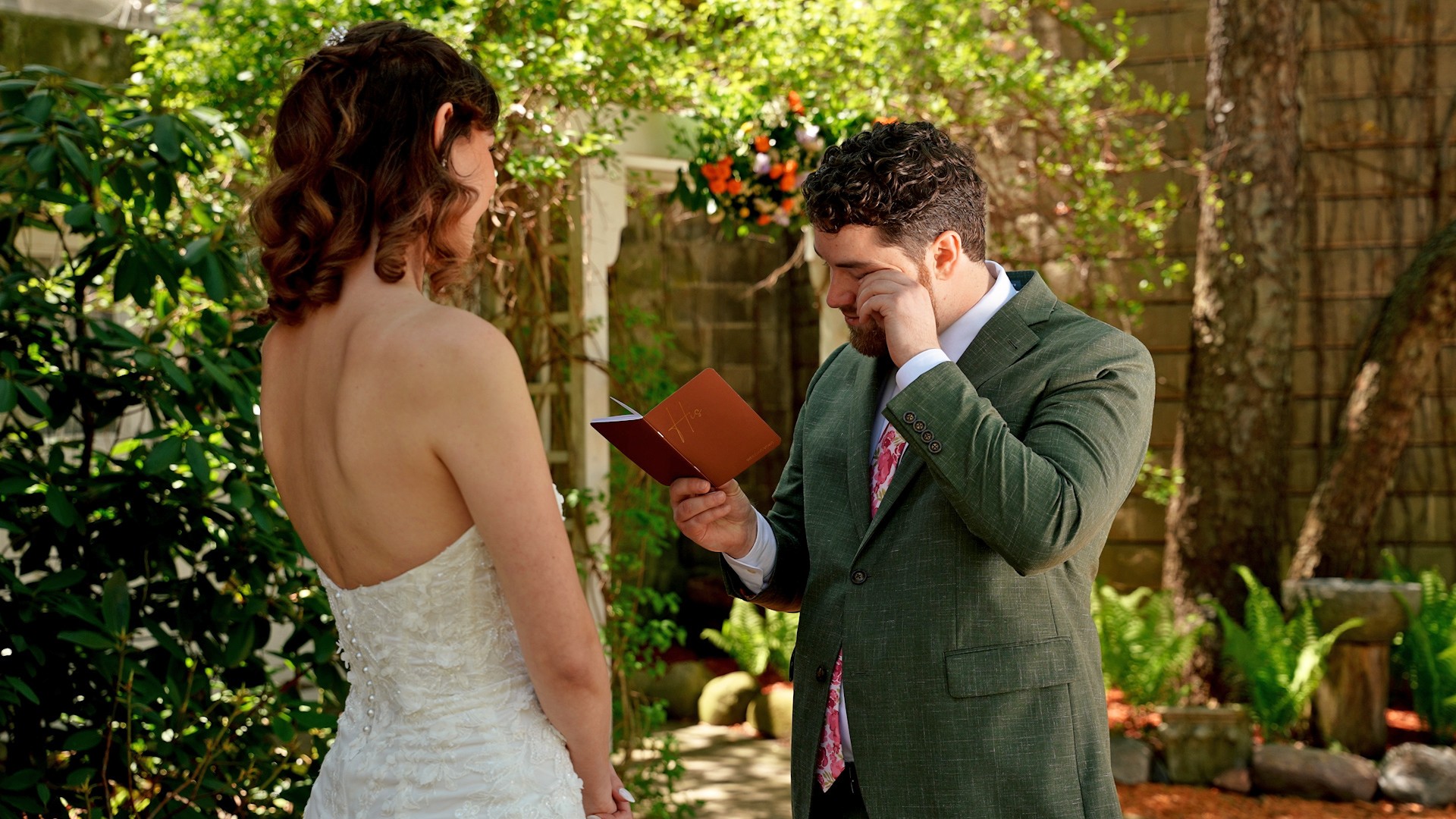 A bride and groom exchanging vows outdoors in a garden setting with greenery and flowers.