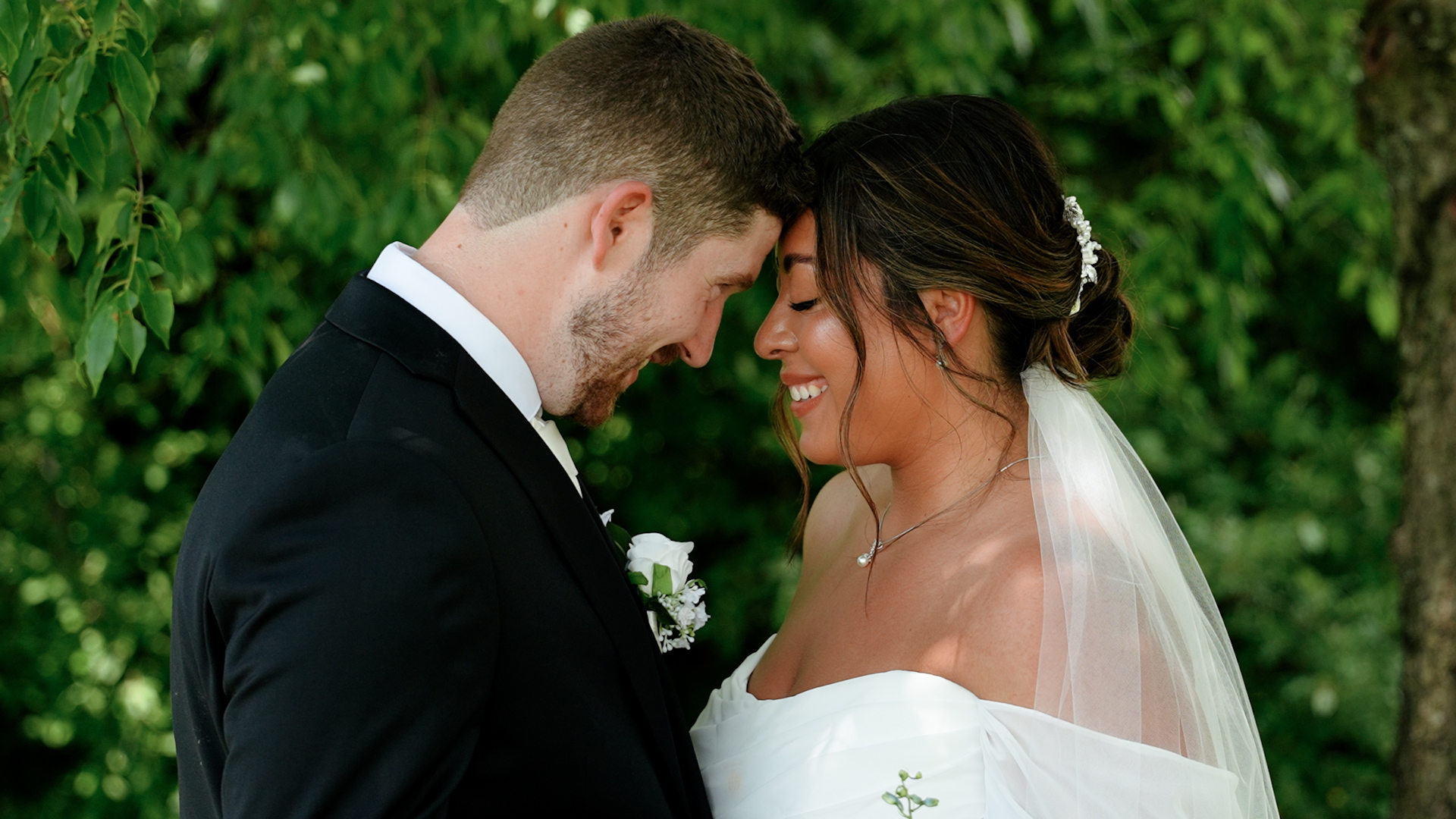 Bride and groom face each other outdoors, smiling, with greenery in the background.