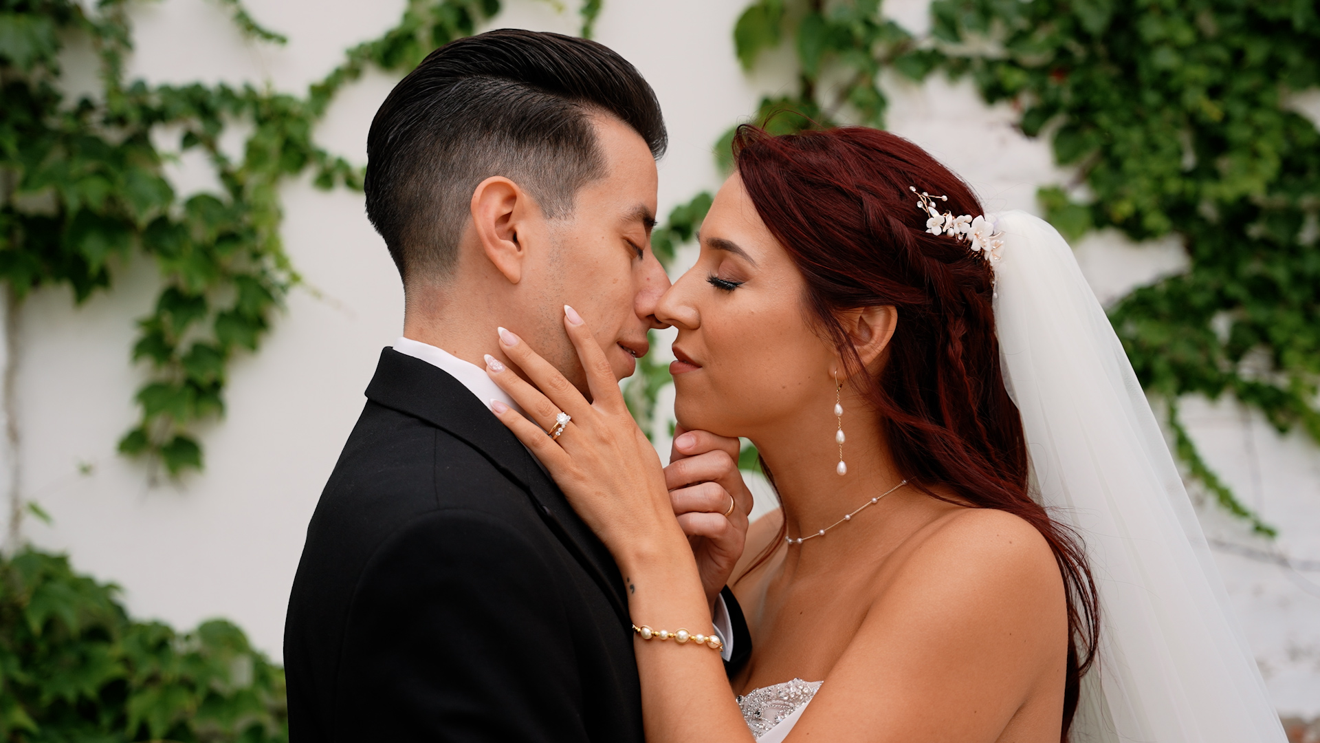 Bride and groom face each other closely, touching noses, outdoors with green foliage in background.