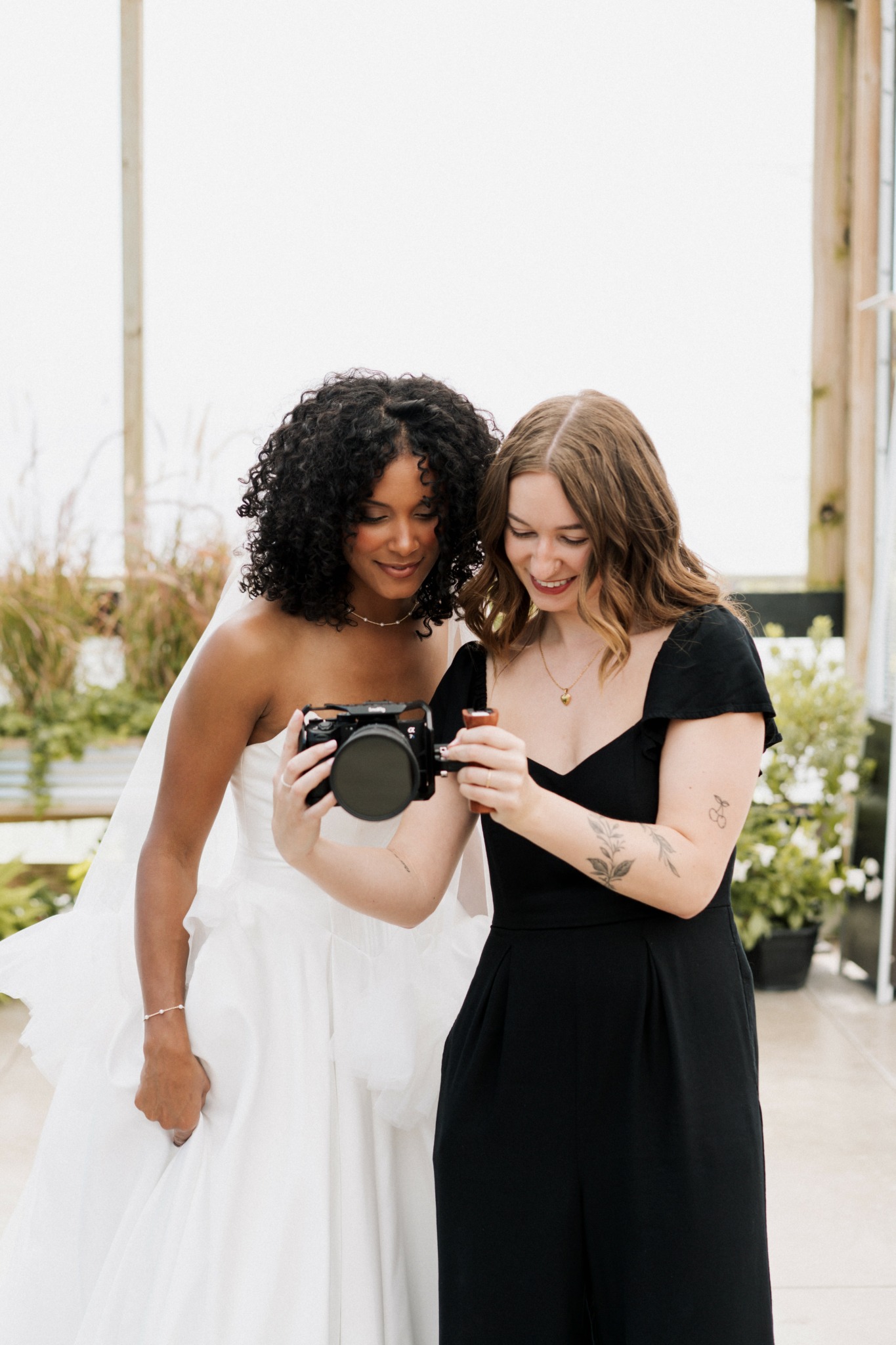 Two women stand together indoors, looking at a camera and a microphone, smiling. One wears a white dress, the other a black dress.