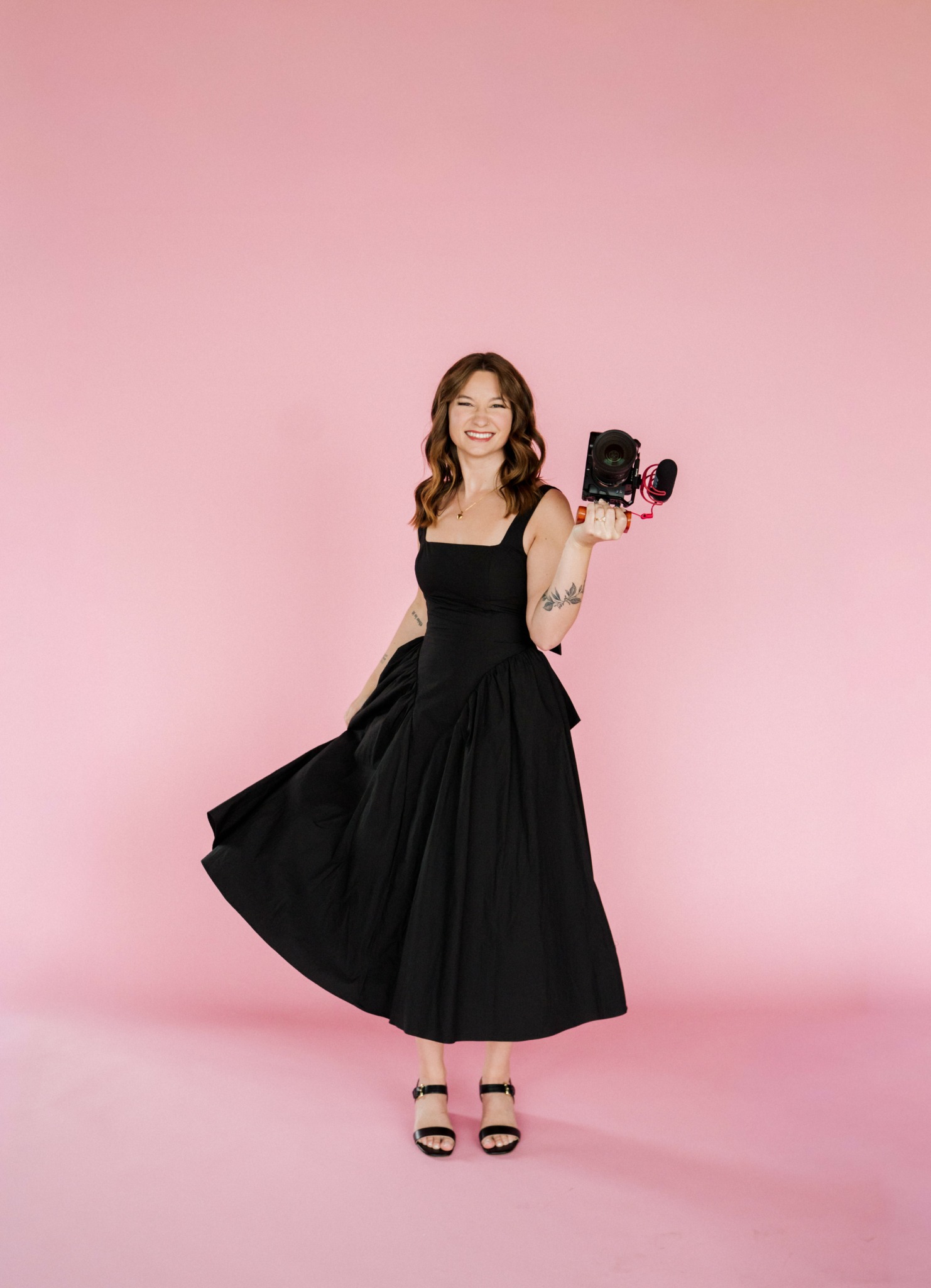 Woman in black dress holding a camera, standing on pink background, smiling, wearing high heels.