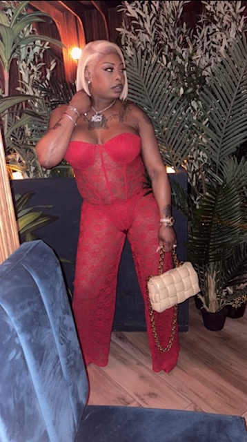 Woman in red lace outfit holding a beige handbag, standing indoors with plants and wooden floor.