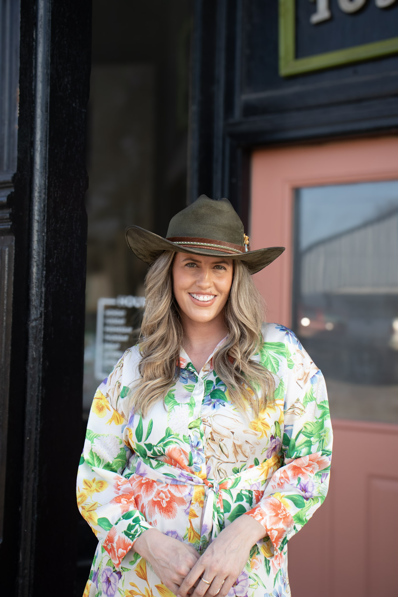 Woman wearing a wide-brimmed hat and a colorful floral shirt, smiling outdoors near a building with pink and blue doors.