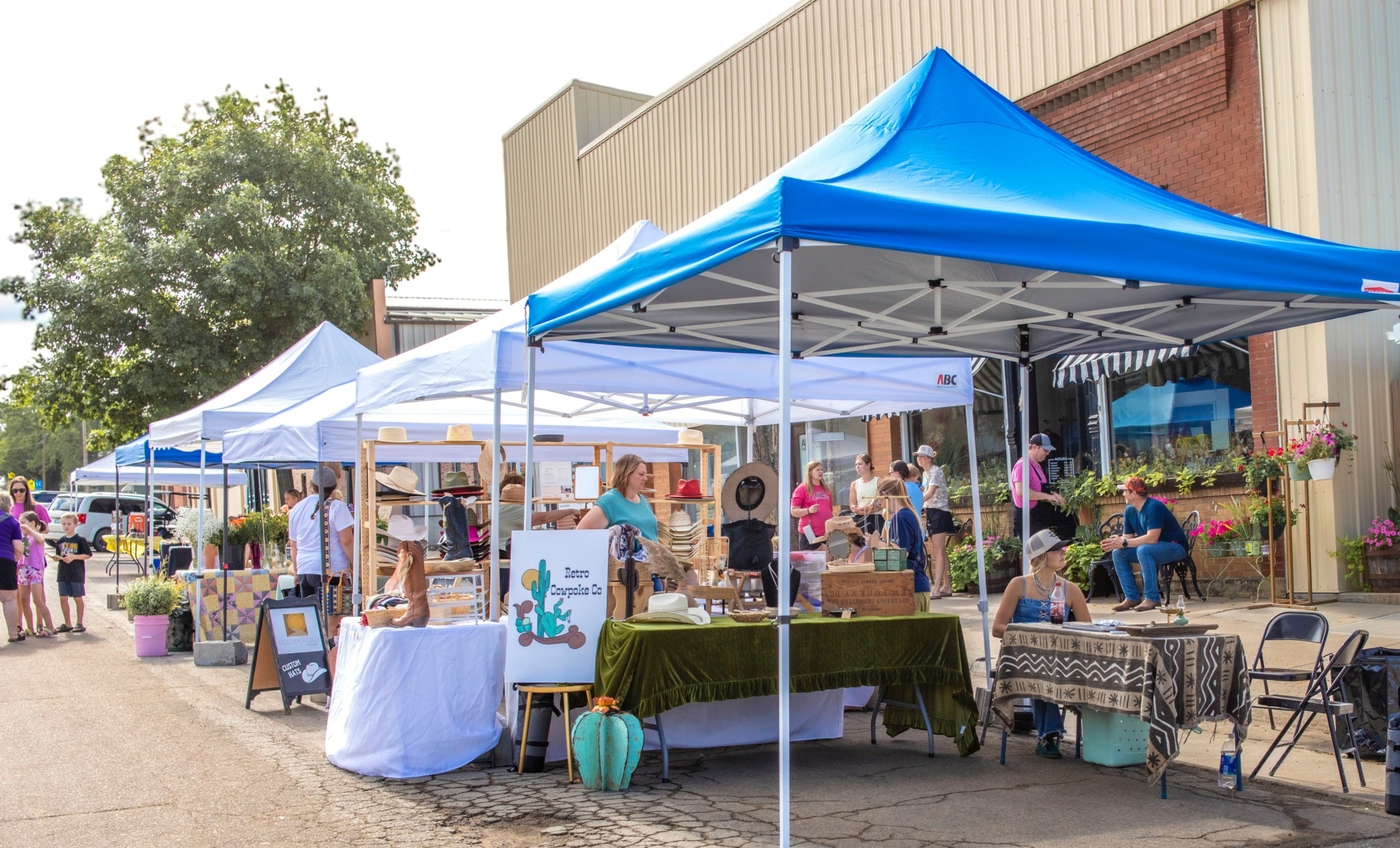 Outdoor market with tents, tables, and people browsing and shopping, set against buildings and trees.