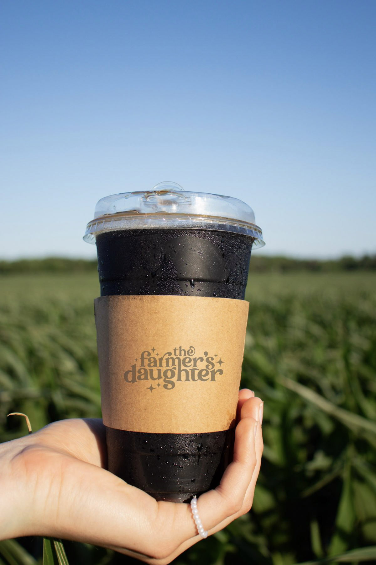 Hand holding a black coffee cup with a brown sleeve outdoors in a green field under a blue sky.