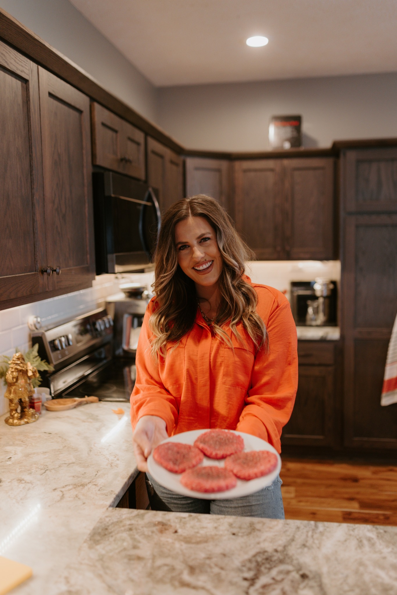 Woman with long hair in orange shirt holding a plate of cupcakes in a kitchen.