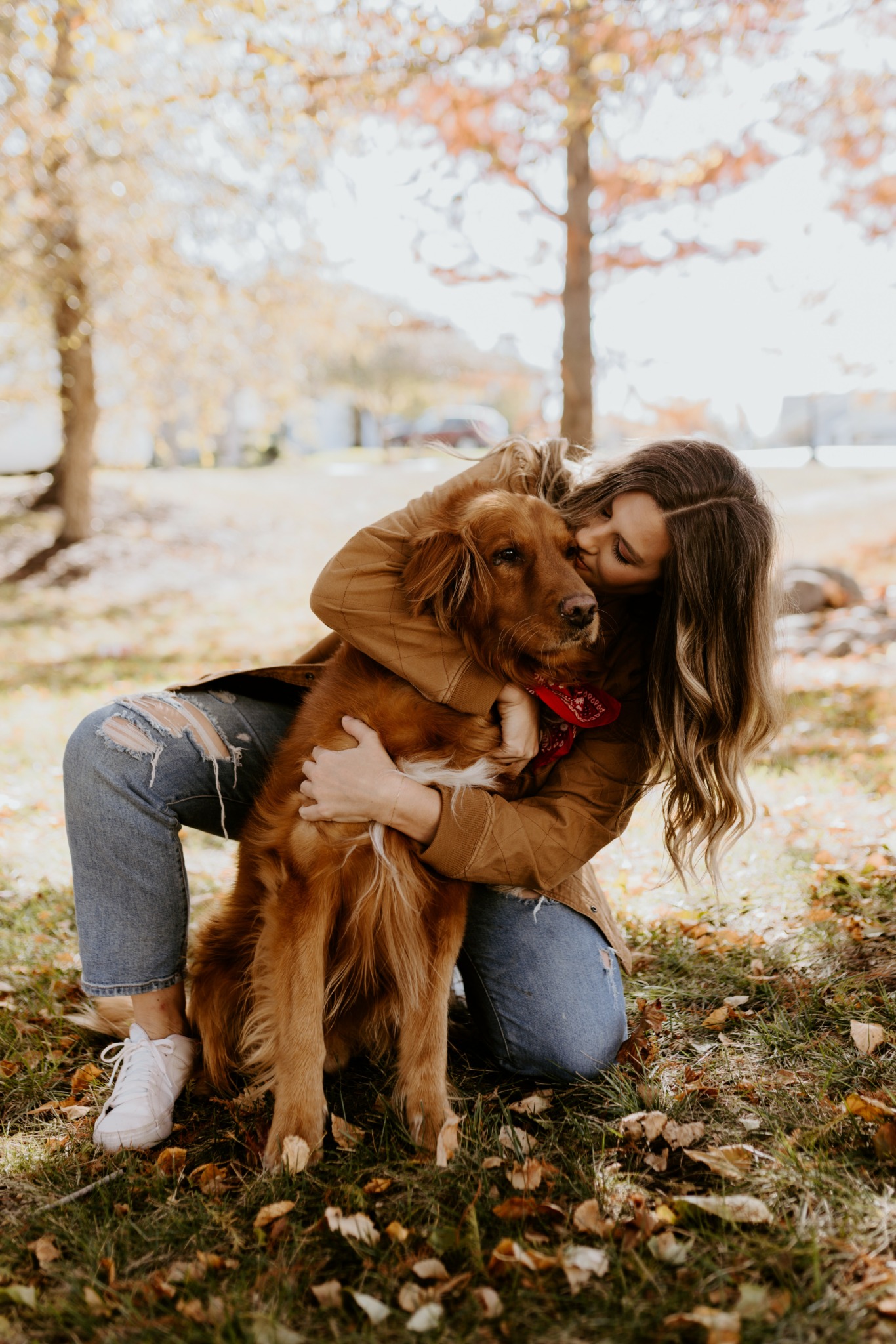 Woman hugging a large dog outdoors in autumn, surrounded by fallen leaves and trees with orange and yellow foliage.