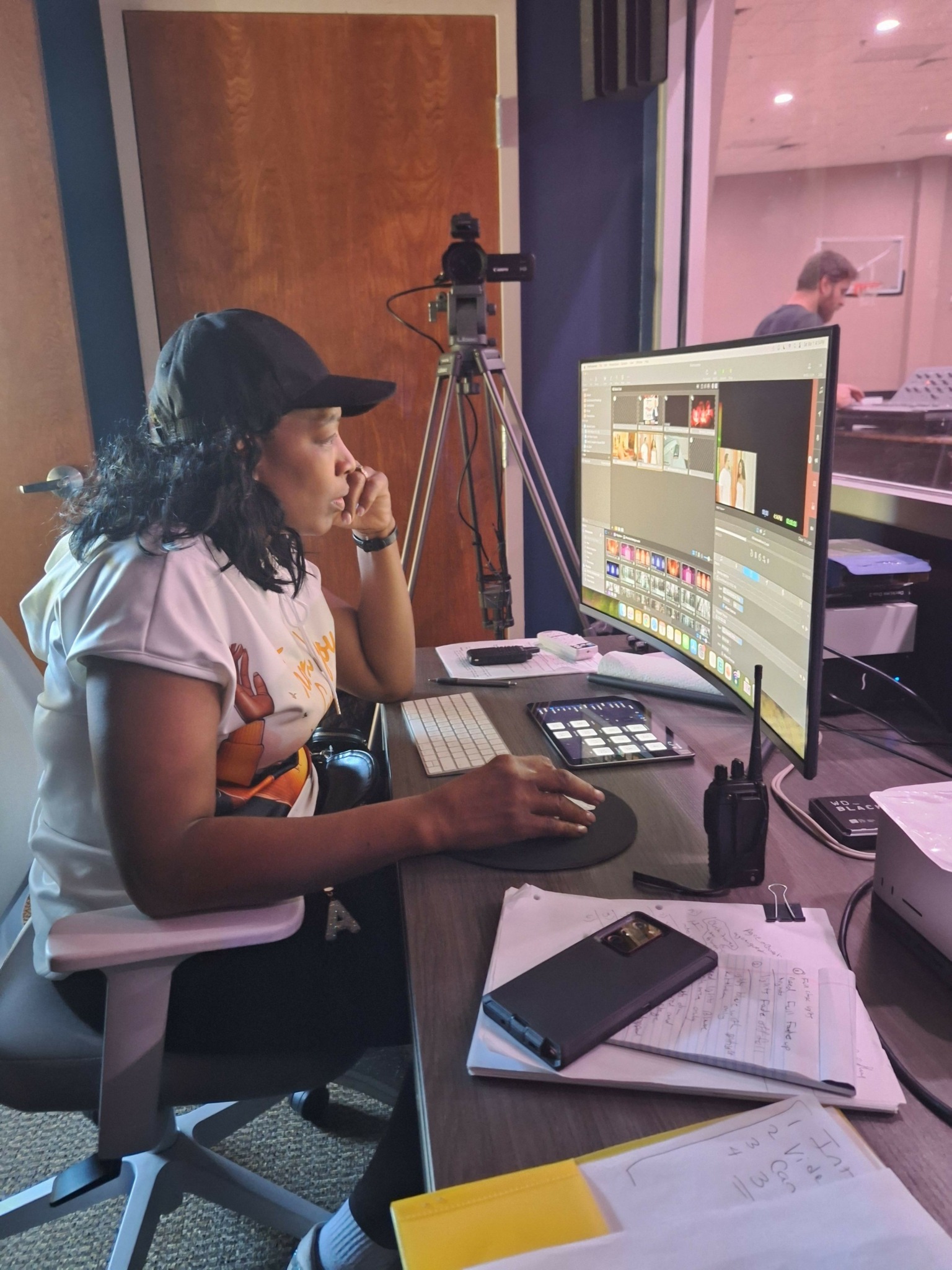Woman working at a computer with editing software, surrounded by notebooks, a phone, and a camera on a tripod.