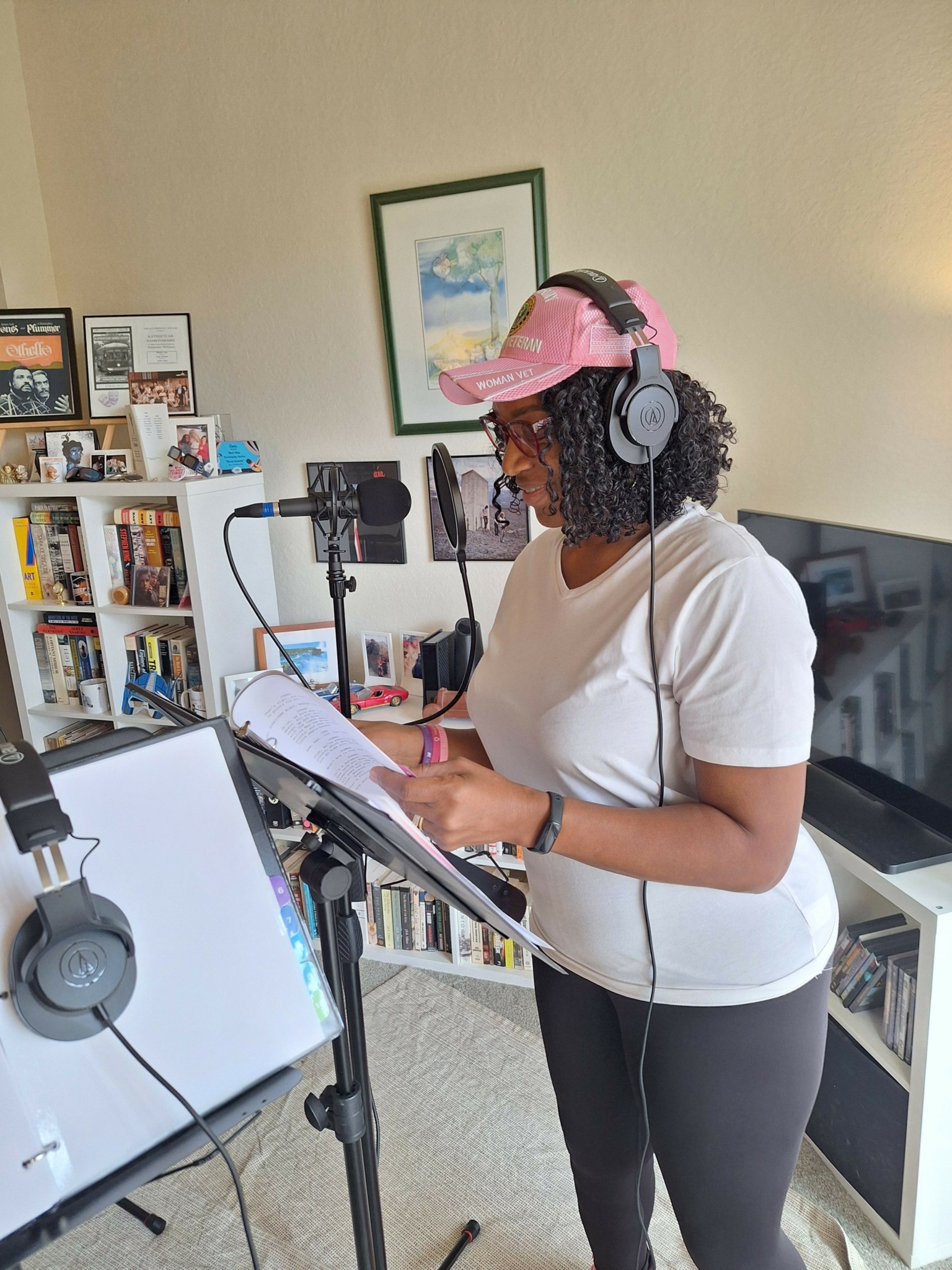 Woman with curly hair wearing pink cap and headphones, reading from a notebook in a room with bookshelves and a TV.