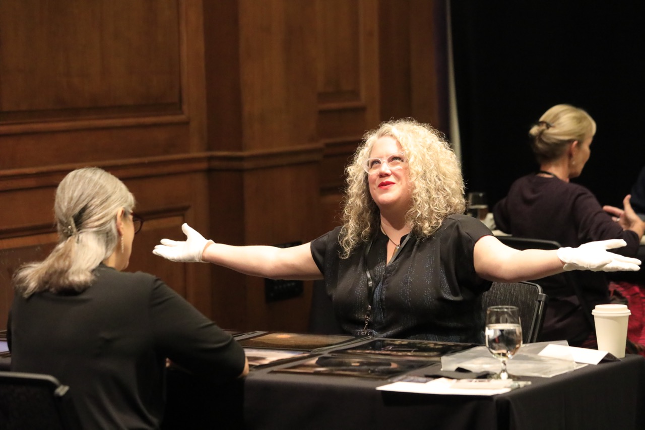 Woman with curly blonde hair and glasses speaking with arms outstretched at a table in a wood-paneled room.