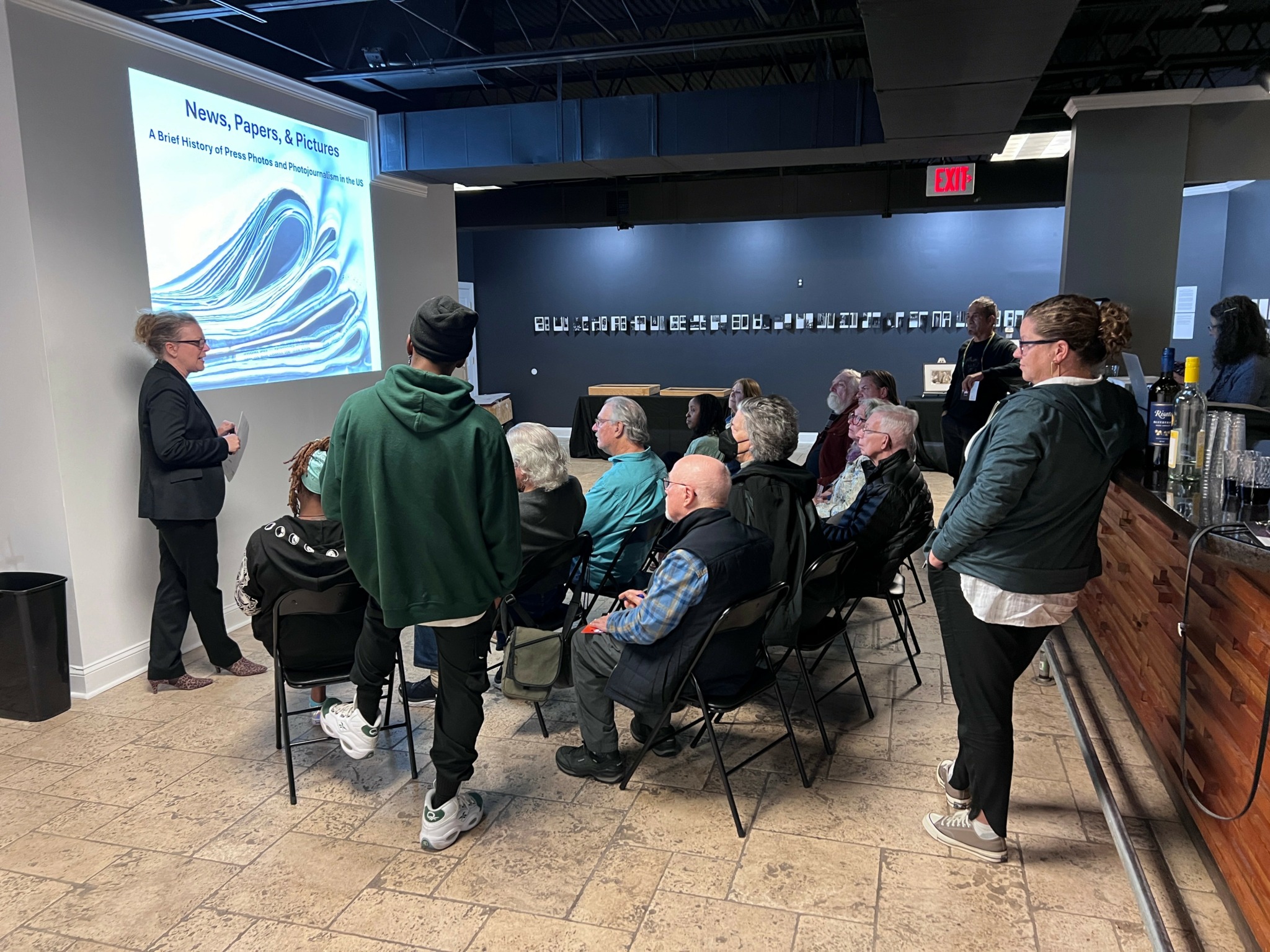 Group of people seated and standing in a room watching a presentation on a screen, with a speaker at the front.