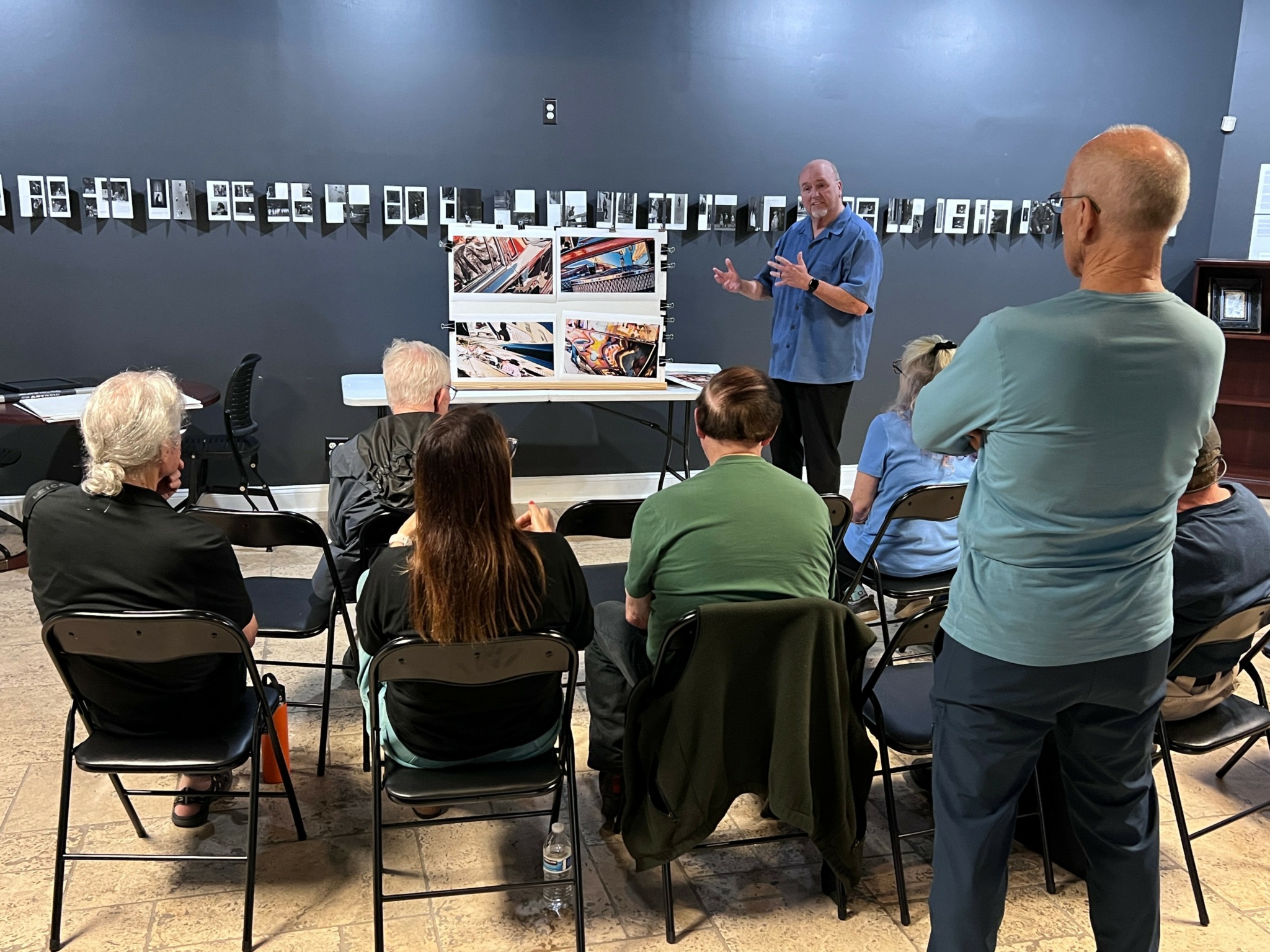 Man presenting to a seated group in an art gallery with photographs on the wall.
