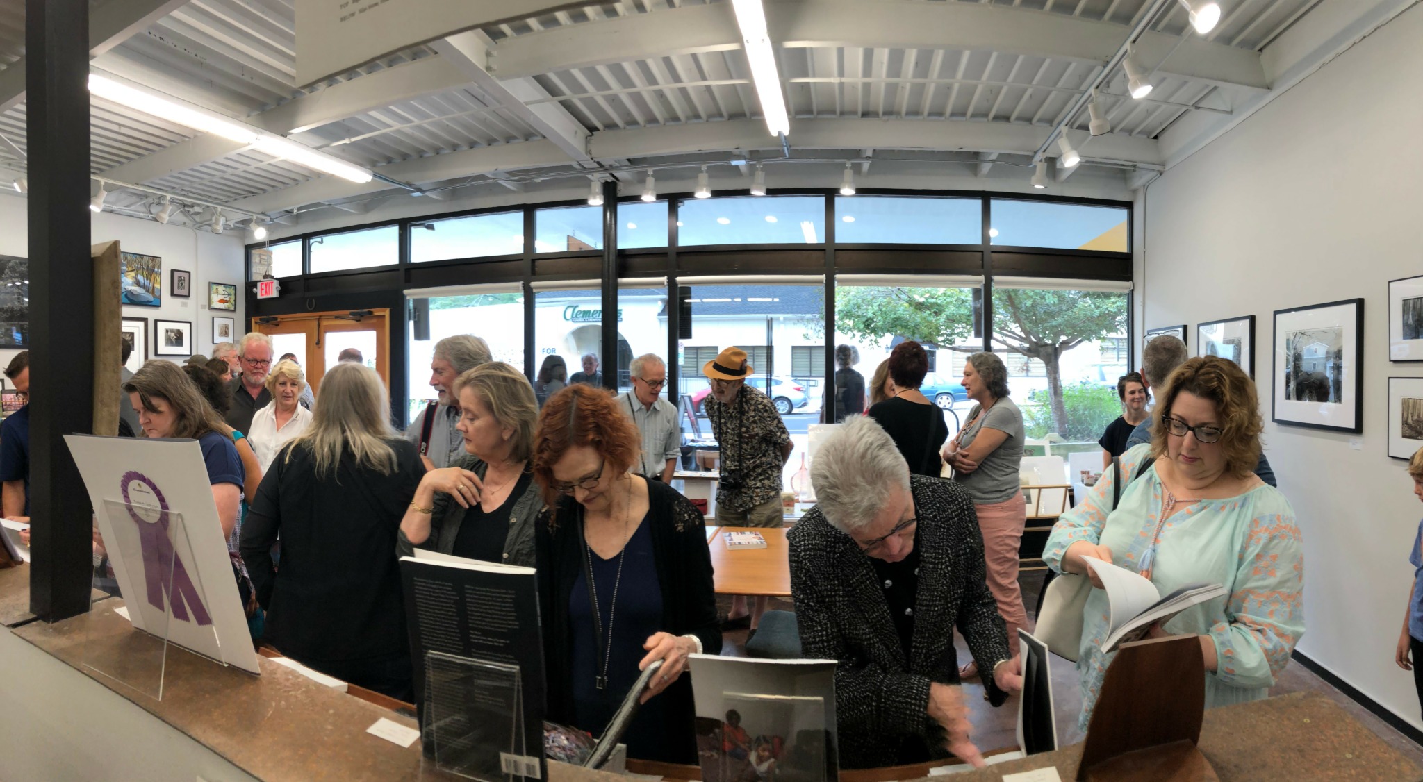 People browsing art gallery with framed pictures on white walls and large windows in background.