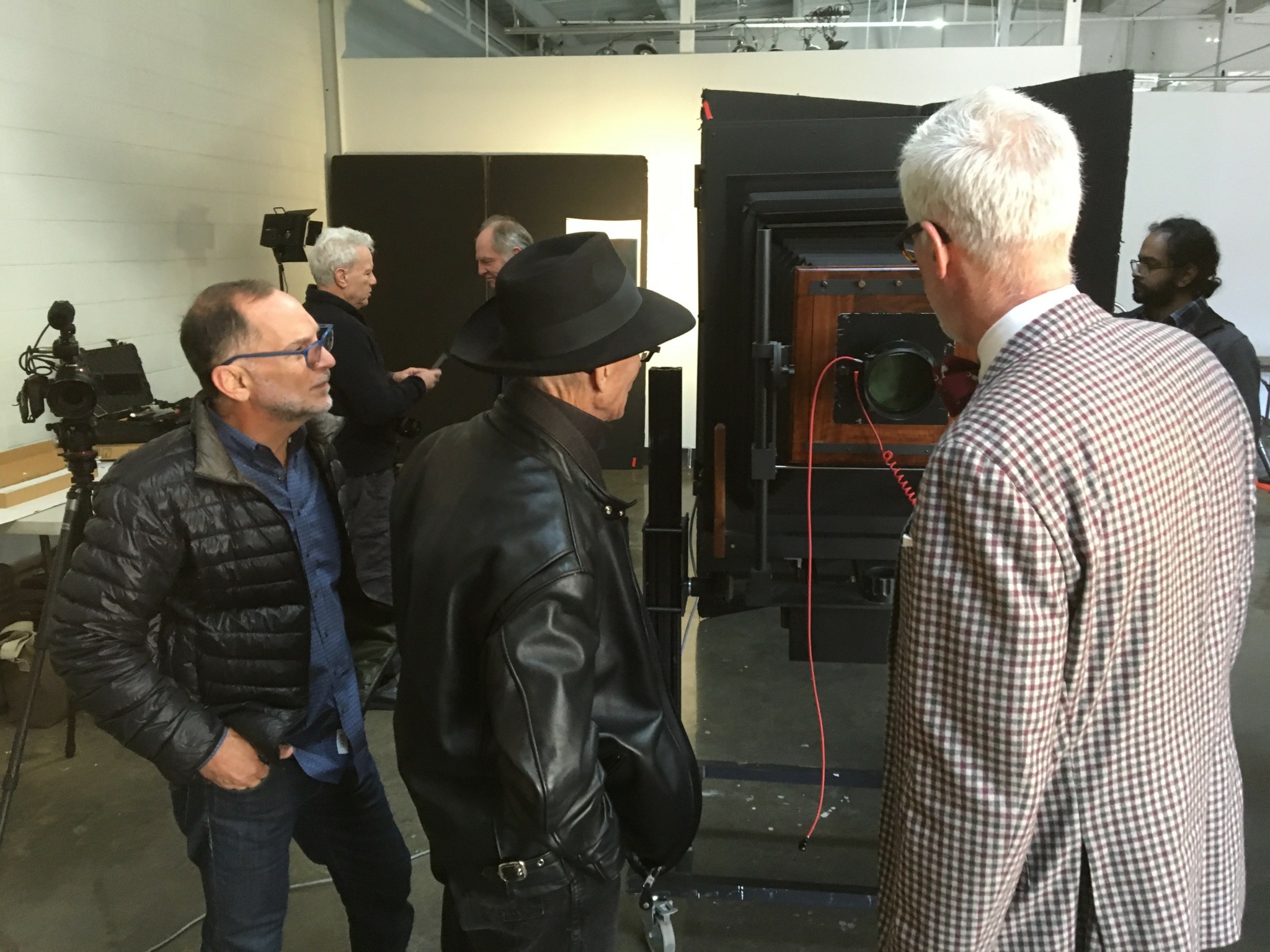 Three people in conversation near a large black machine at an indoor event, with cameras and other people in background.