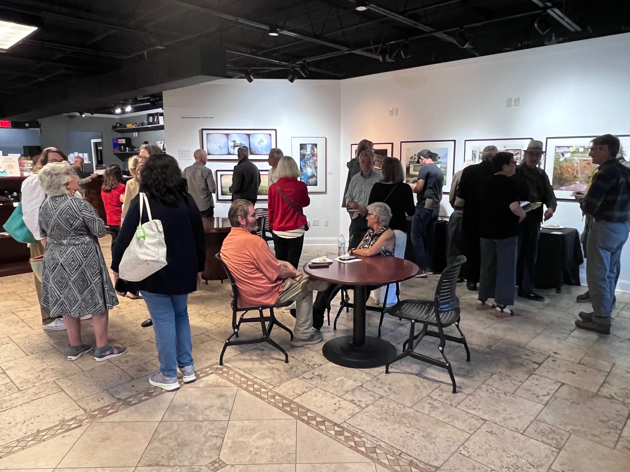 People viewing artwork in an art gallery, some standing and some sitting at tables, with framed art on white walls.