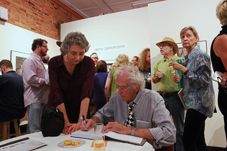 Group of people gathered indoors, some talking, some reading, with a white wall and brick wall in background.