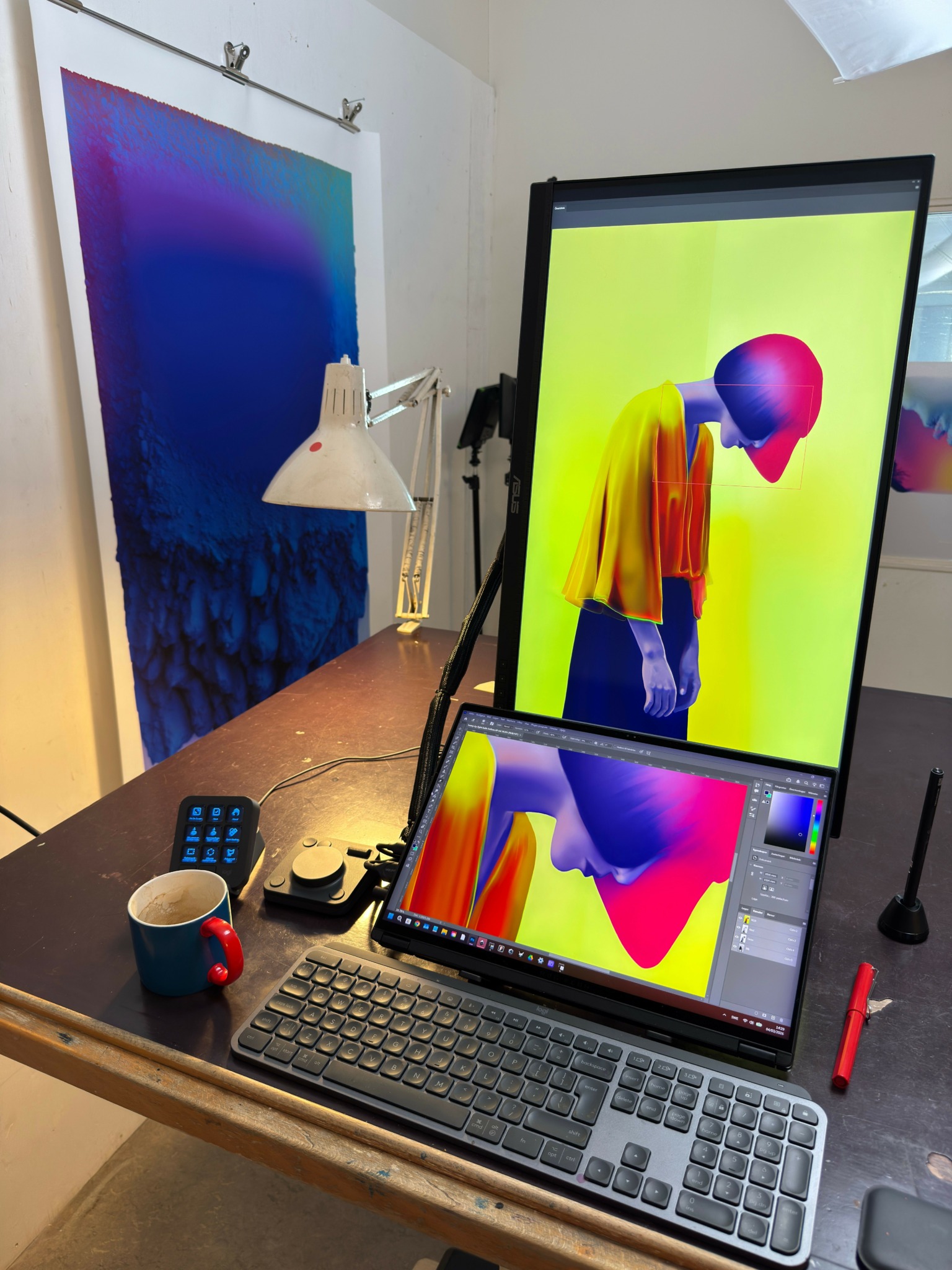 Desk with a laptop, monitor, keyboard, mug, and desk lamp in a room with artwork on the wall.