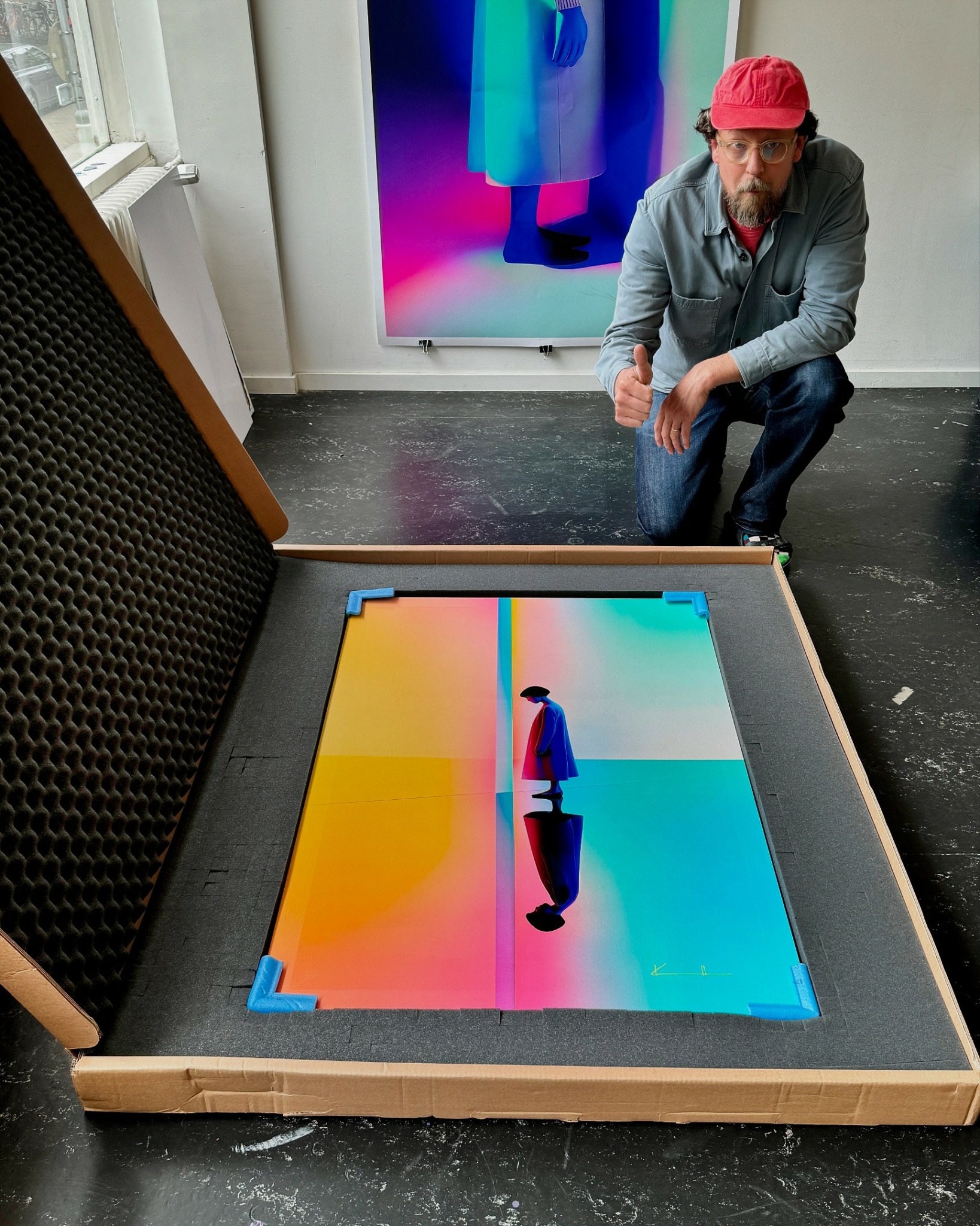 Man with a red cap crouching near a colorful digital artwork on the floor inside a display case.