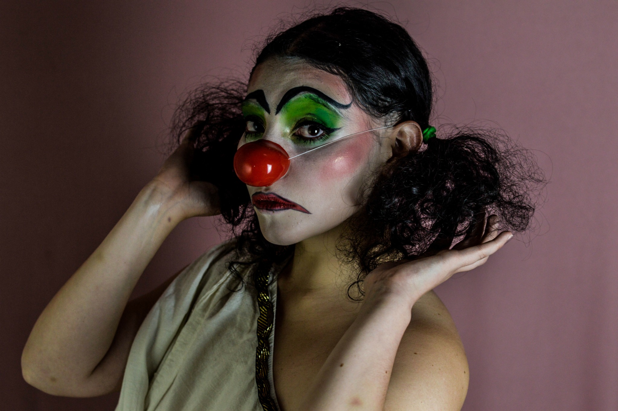 Person with clown makeup, green eye shadow, red nose, and curly hair, posing with hands near face.