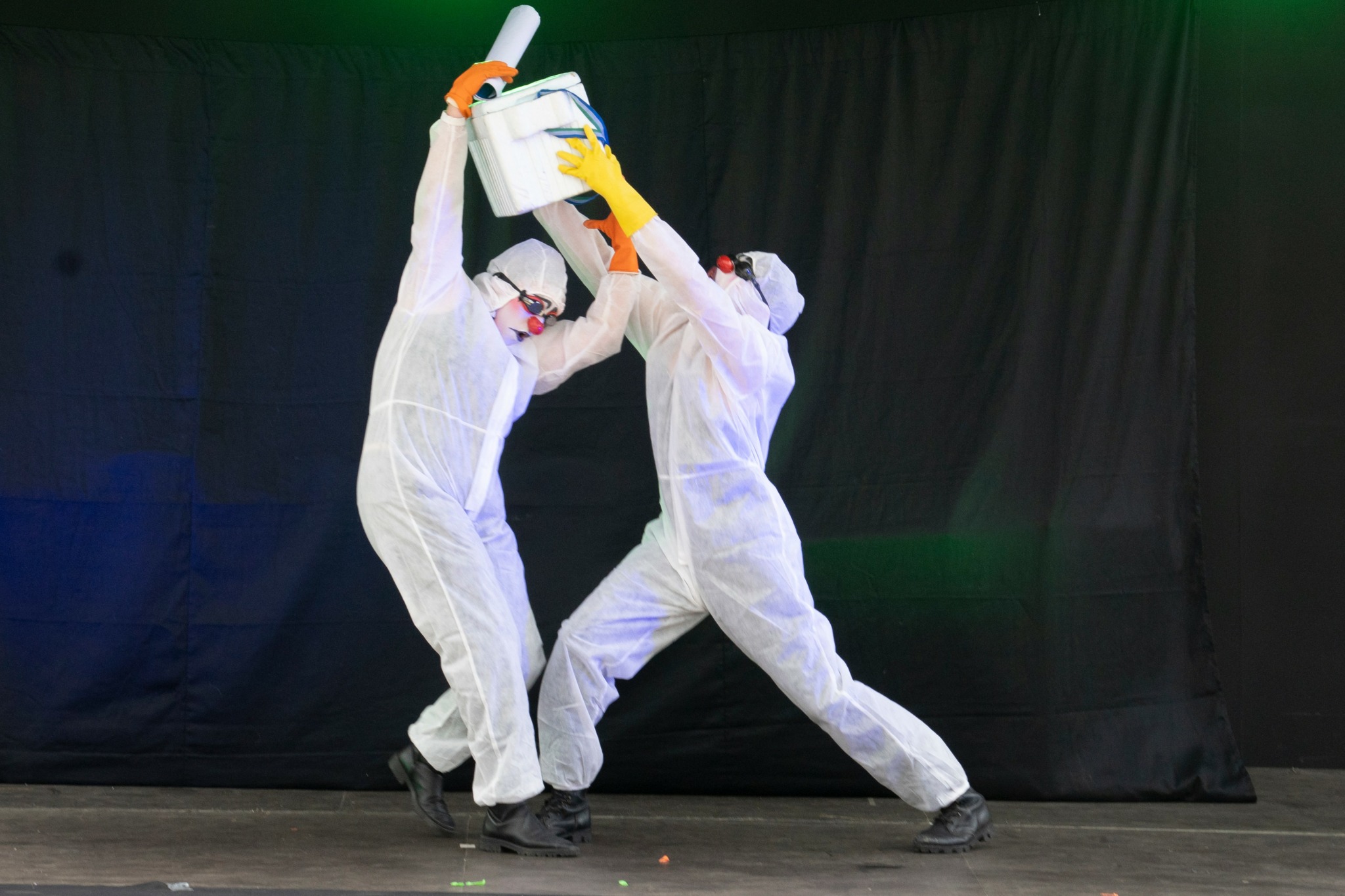 Two people in white protective suits and yellow gloves hold a large container together against a black background.