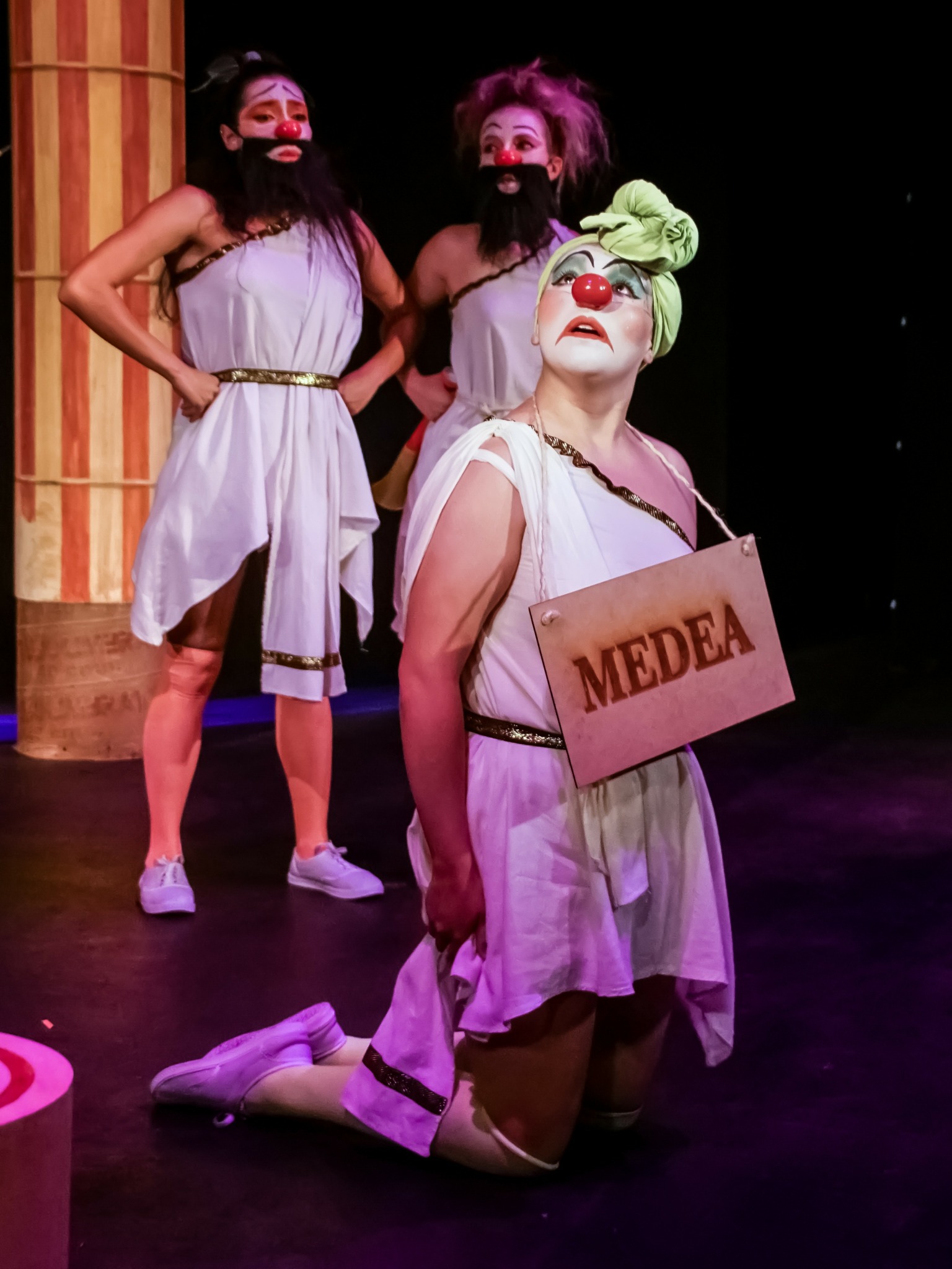 Person dressed as clown with green headwrap and red nose holds a sign reading 'MEDEA' on stage.