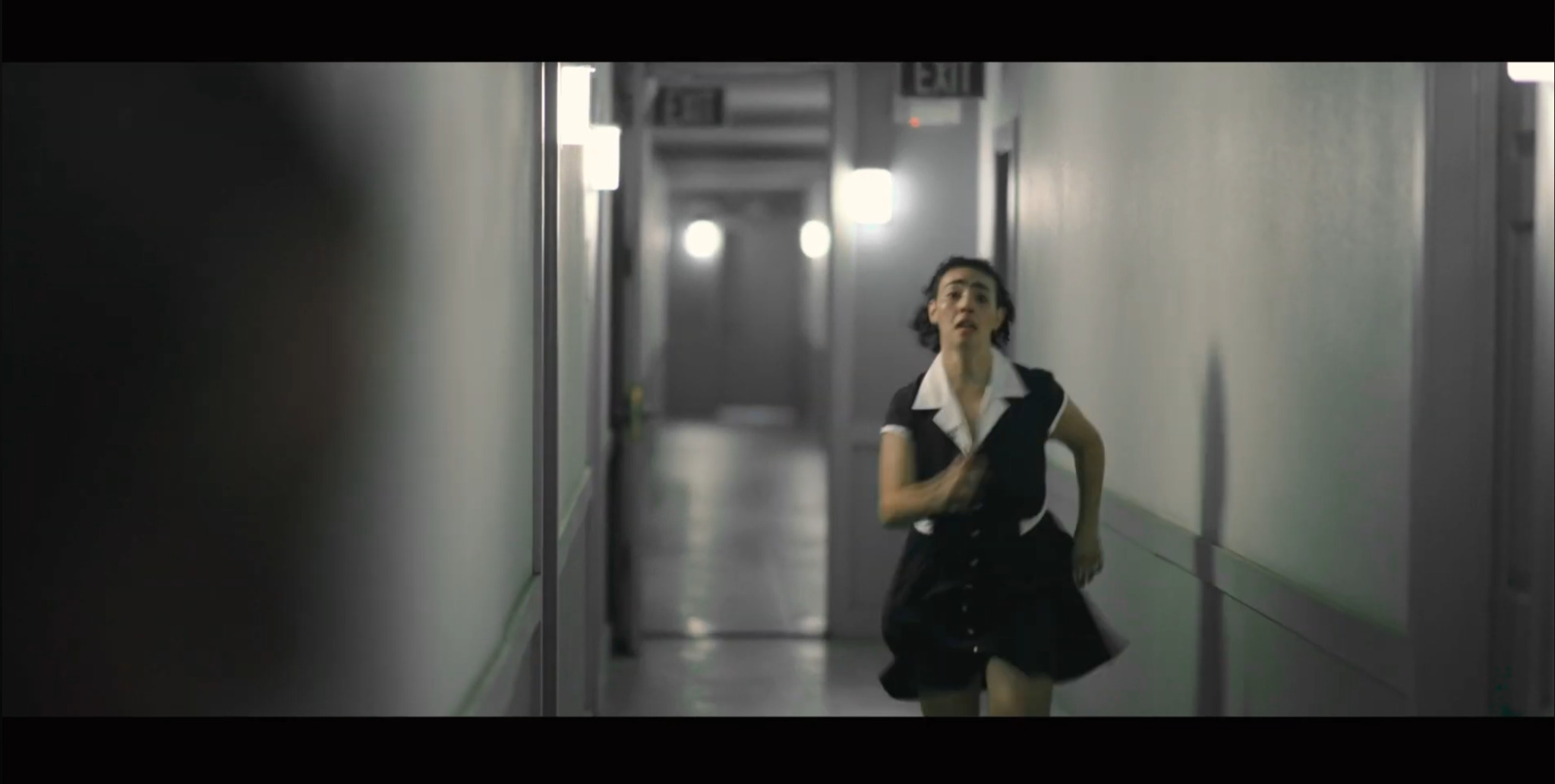 Woman in black dress running down a corridor with plain walls and bright lighting.