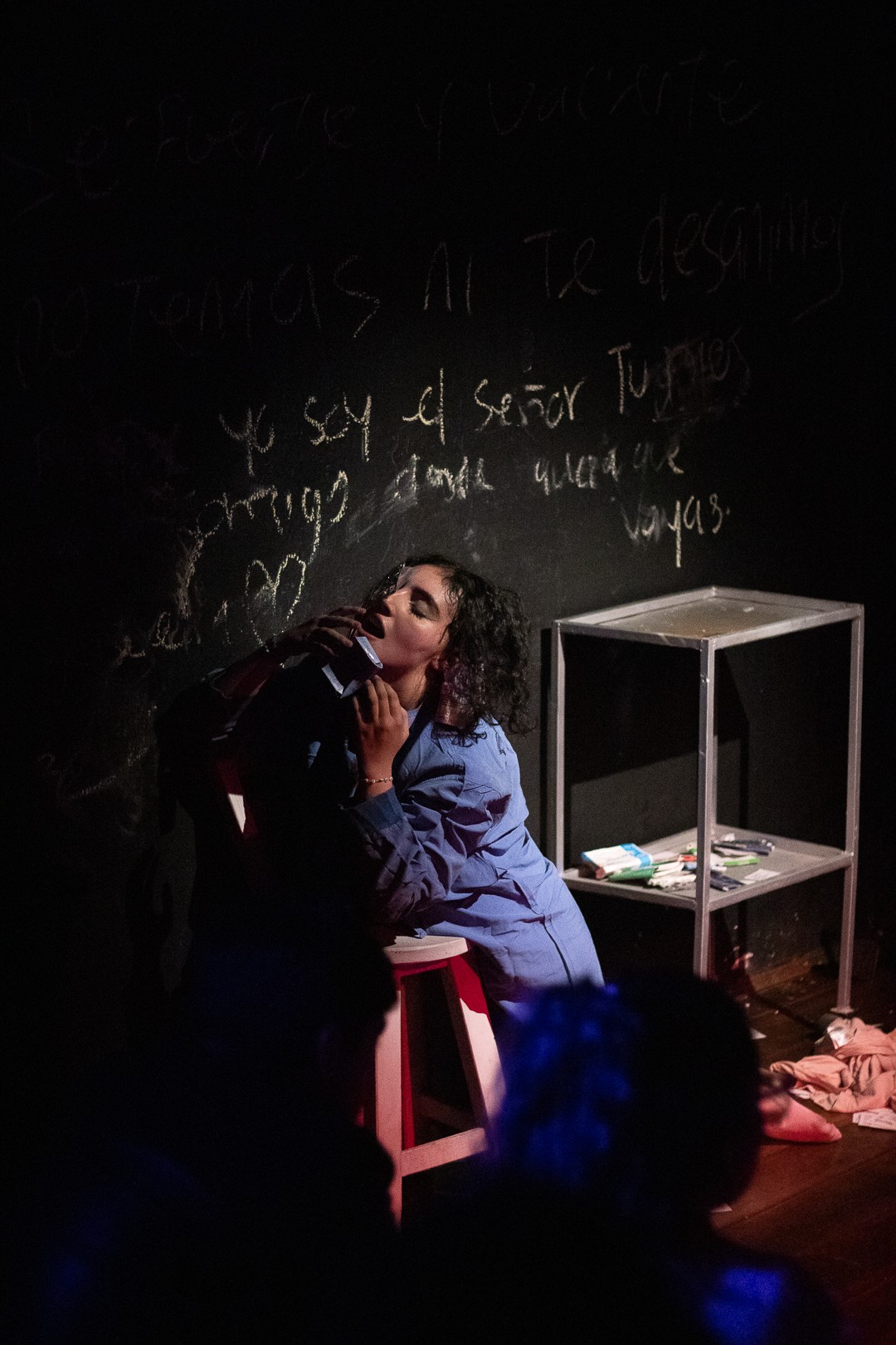 Woman sitting on a stool, leaning on her hand, in a dark room with writing on the wall behind her.