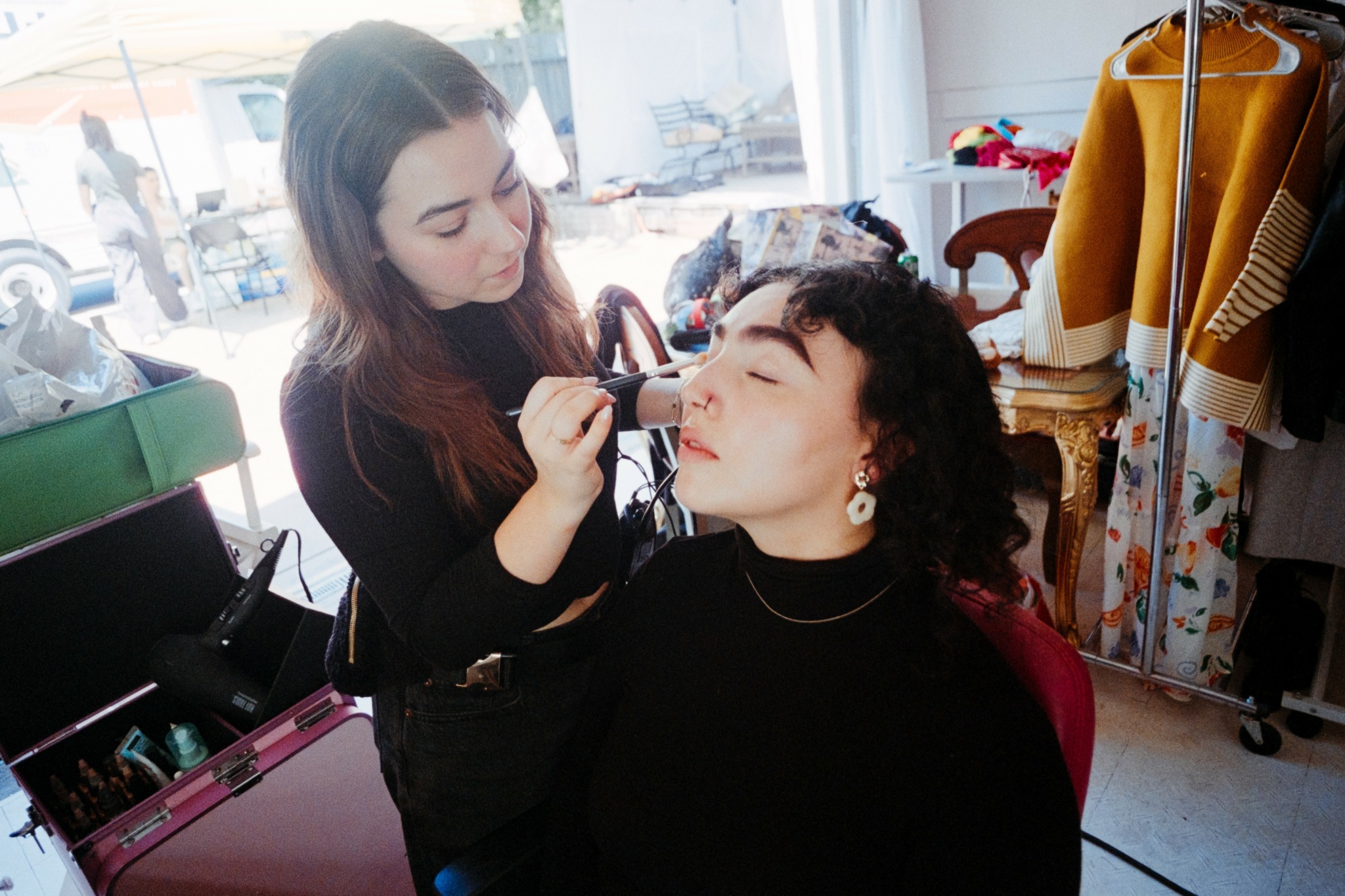 Makeup artist applying makeup to a woman seated in a room with clothing and accessories in the background.