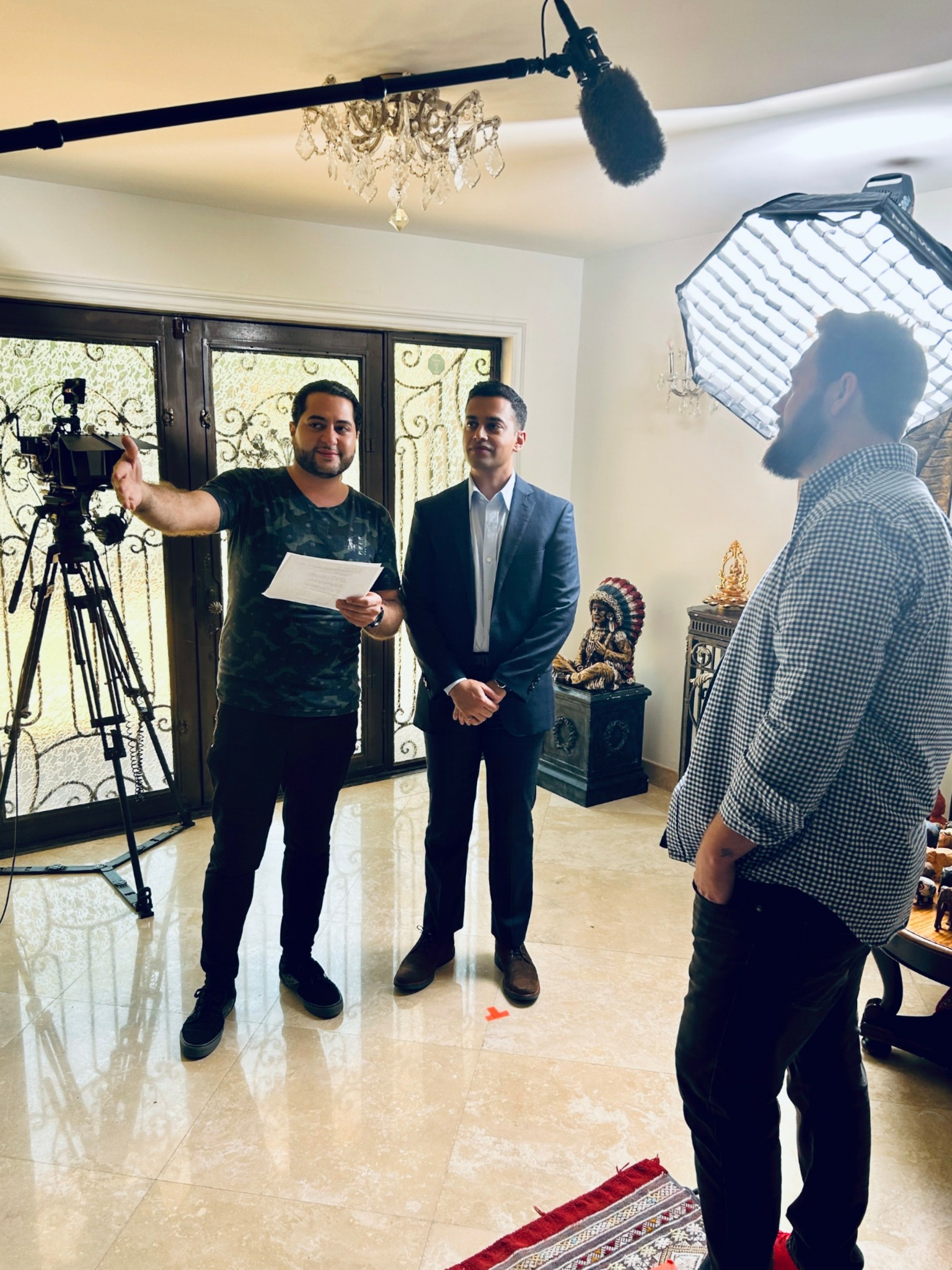 Three men in a room with filming equipment, one holding papers, another in a suit, and the third with a beard and glasses.