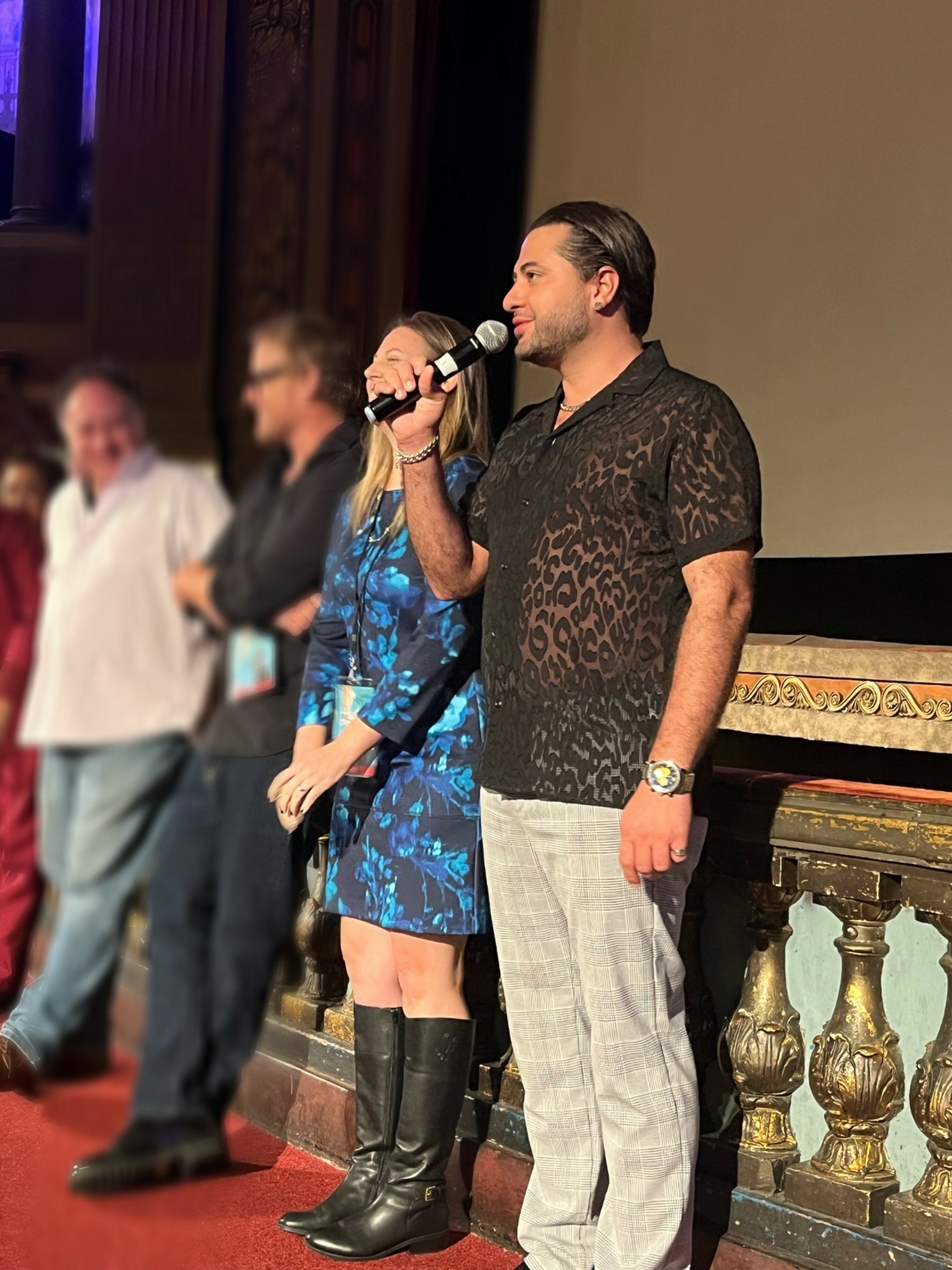 Man holding microphone on stage with people in background, ornate railing, and large screen behind him.