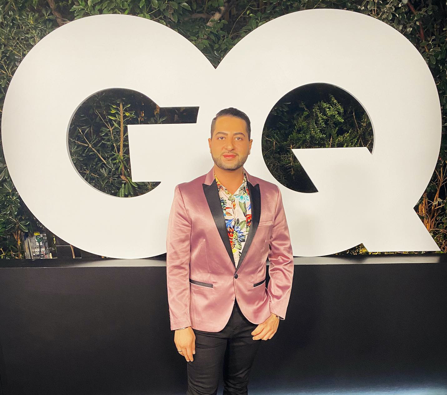Man in pink blazer and floral shirt standing in front of large GQ logo at an outdoor event.