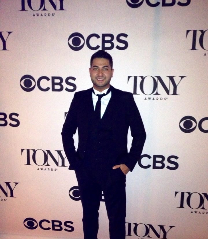 Man in a black suit and tie standing in front of a backdrop with CBS and Tony Awards logos.