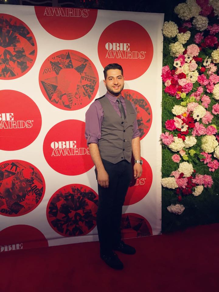 Man in purple shirt and gray vest standing in front of a backdrop with red and white circles and floral arrangement.
