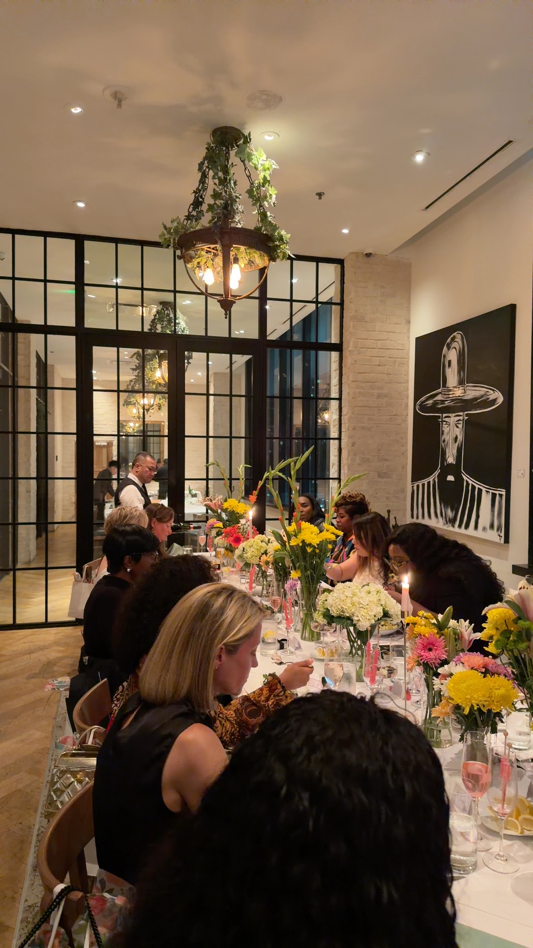 People seated at a table with floral centerpieces in a decorated indoor space with large windows and artwork.