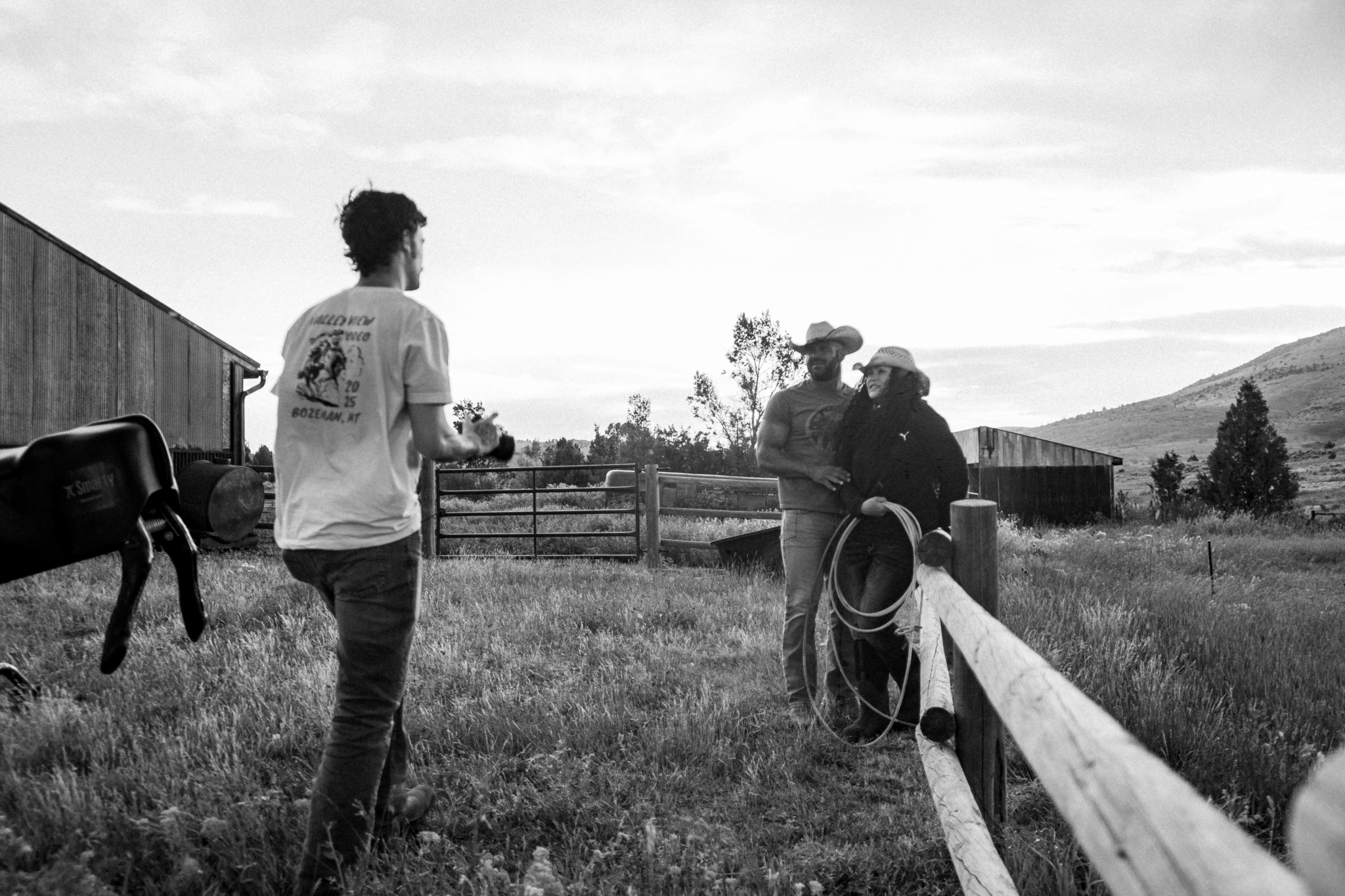Person walking in a grassy field with two people near a fence and a horse nearby, landscape in background.