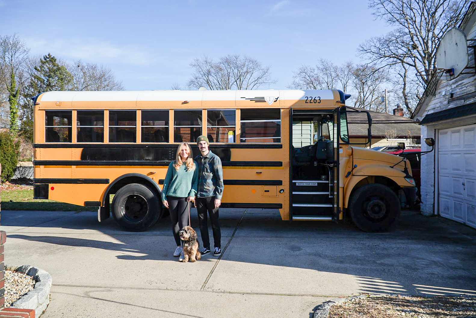 Two people and a dog stand in front of a yellow school bus parked on a driveway.