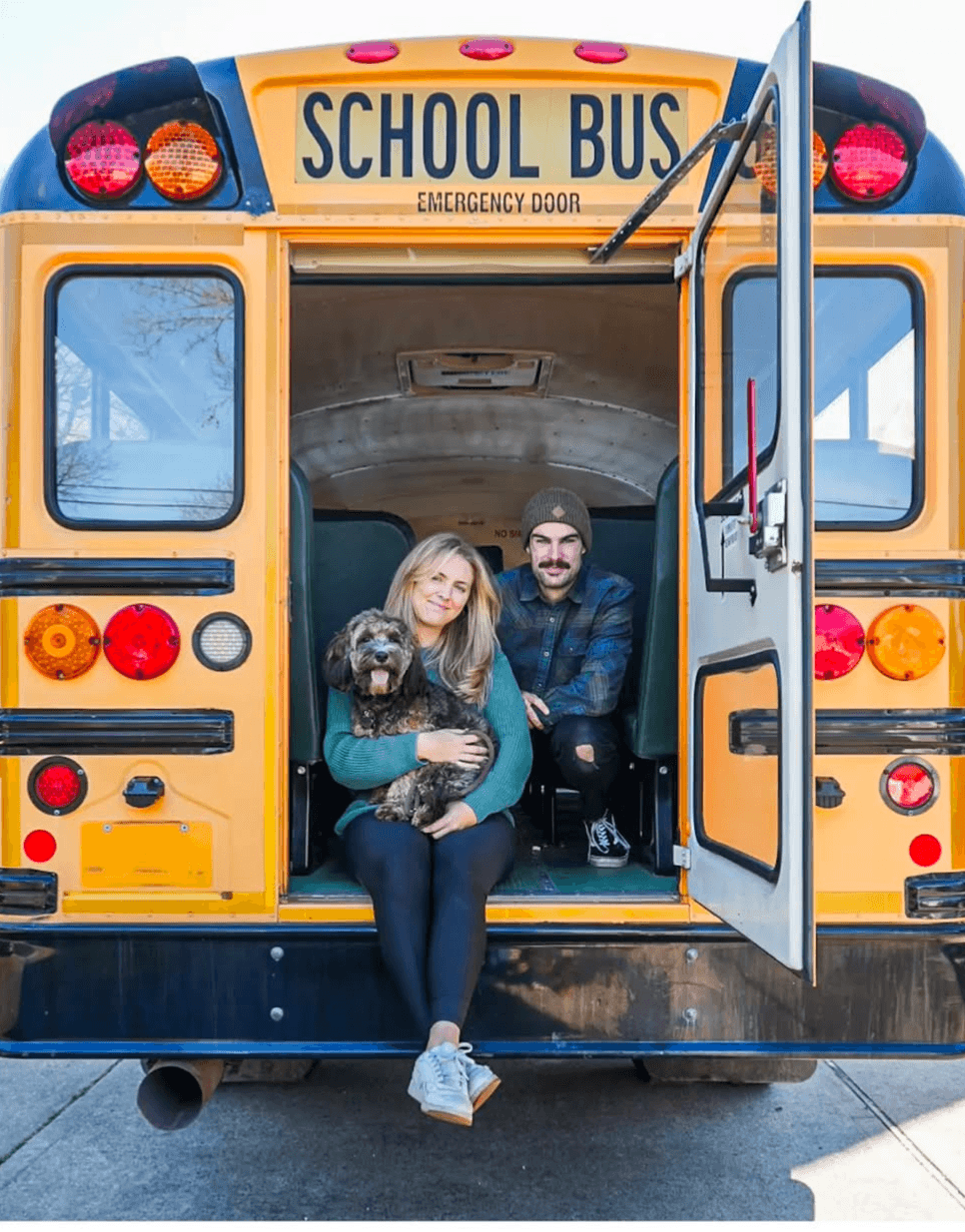 Two people sitting in the open back of a yellow school bus, with a dog on their lap, smiling.