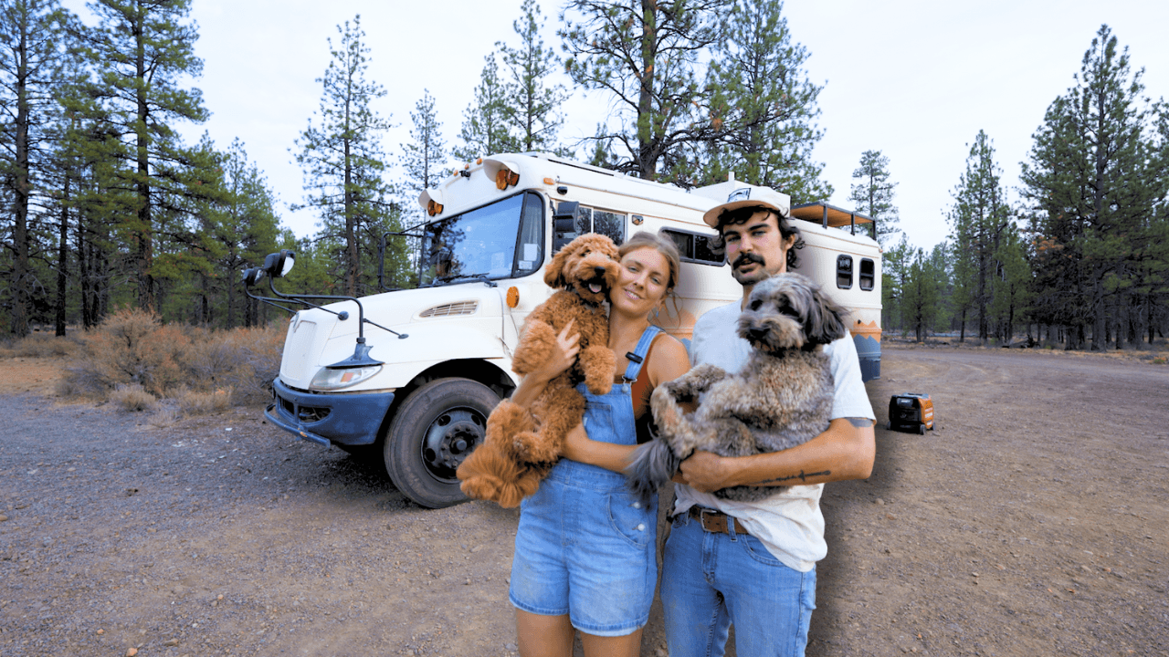 A couple holding dogs stands in front of a white van in a forested area.