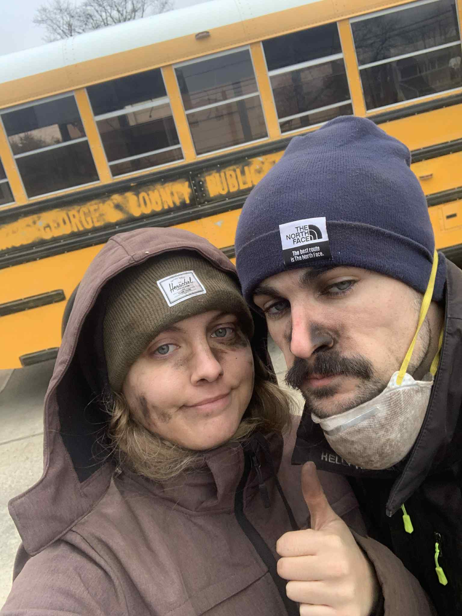Two people in winter clothing take a selfie in front of a yellow school bus.